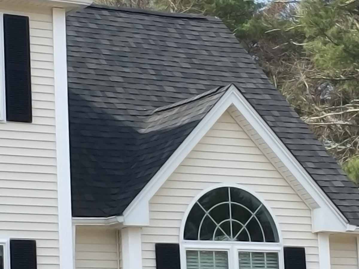 A beige house exterior with dark grey asphalt shingles on the roof and a large, arched window above the siding.