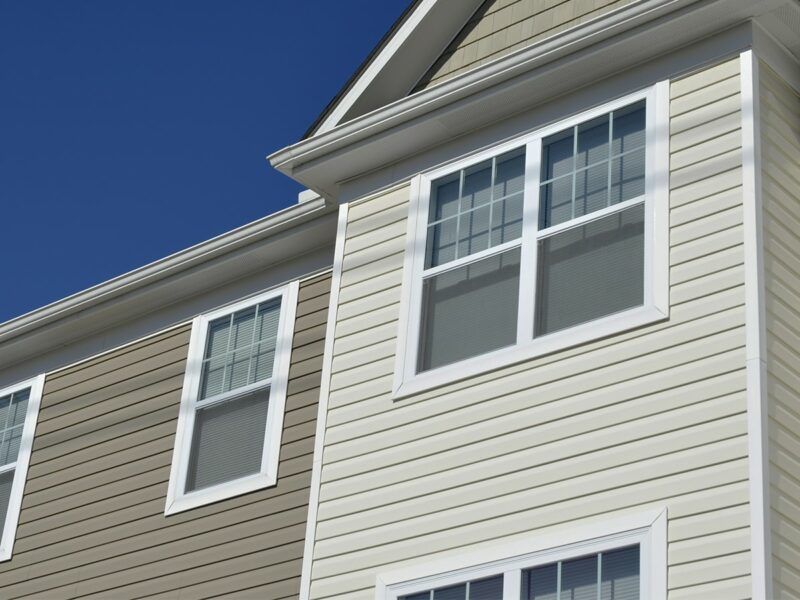 Exterior view of a two-story residential building with beige horizontal siding and white-framed windows against a blue sky.