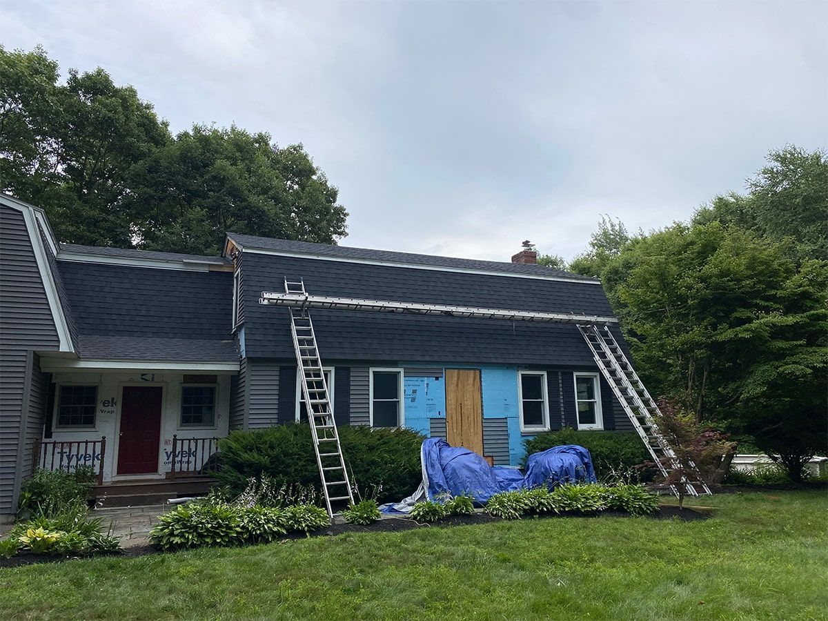 A gray house under renovation with scaffolding and ladders against the roof, blue insulation panels, and bushes in front.