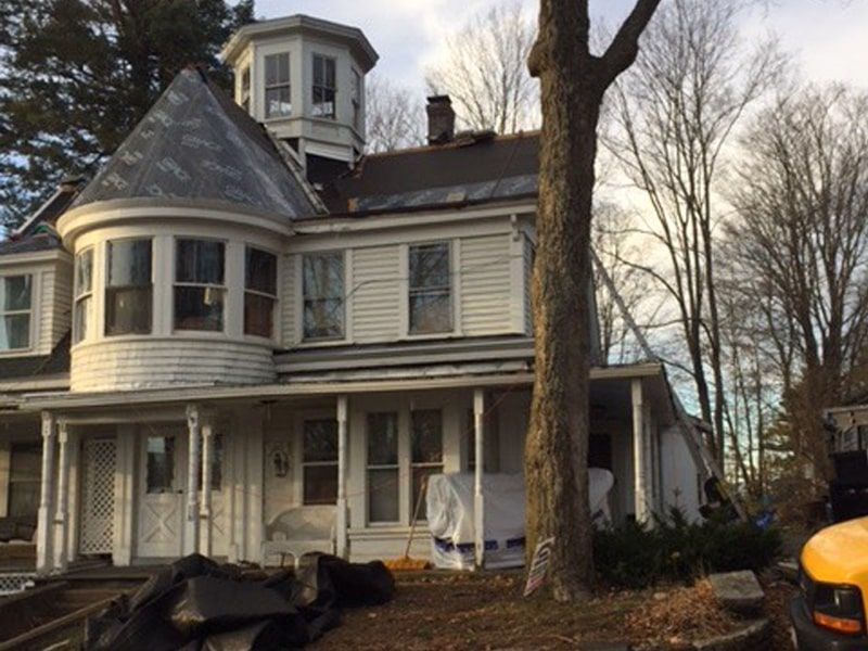 A two-story, white, Victorian-style house undergoing exterior renovations with a tall turret and a large tree in the yard.