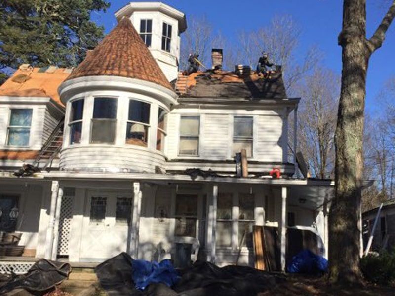 Workers on the roof of a two-story house with a turret and white siding during a renovation project.