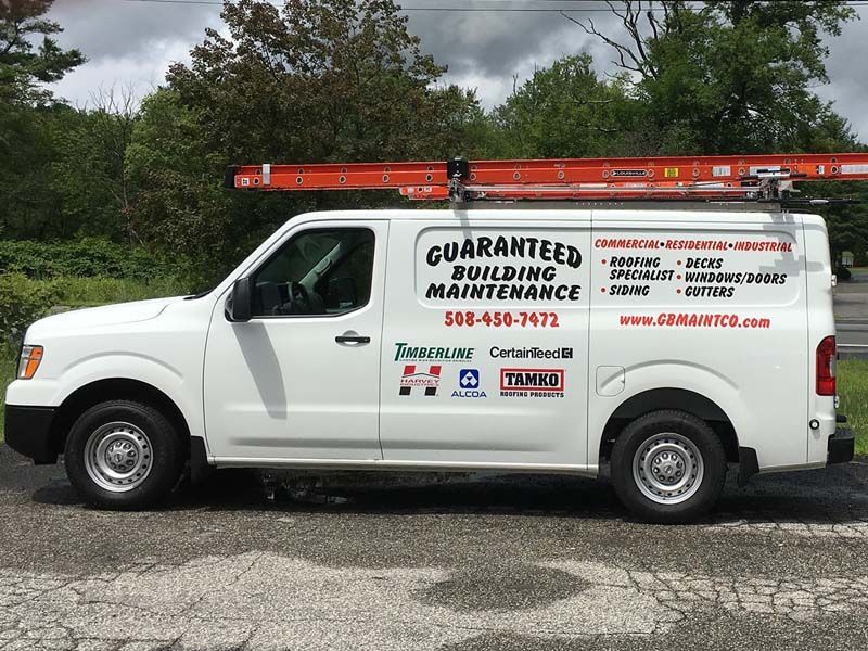 A white Guaranteed Building Maintenance work van with a ladder on the roof, parked on a paved lot with trees in the back.