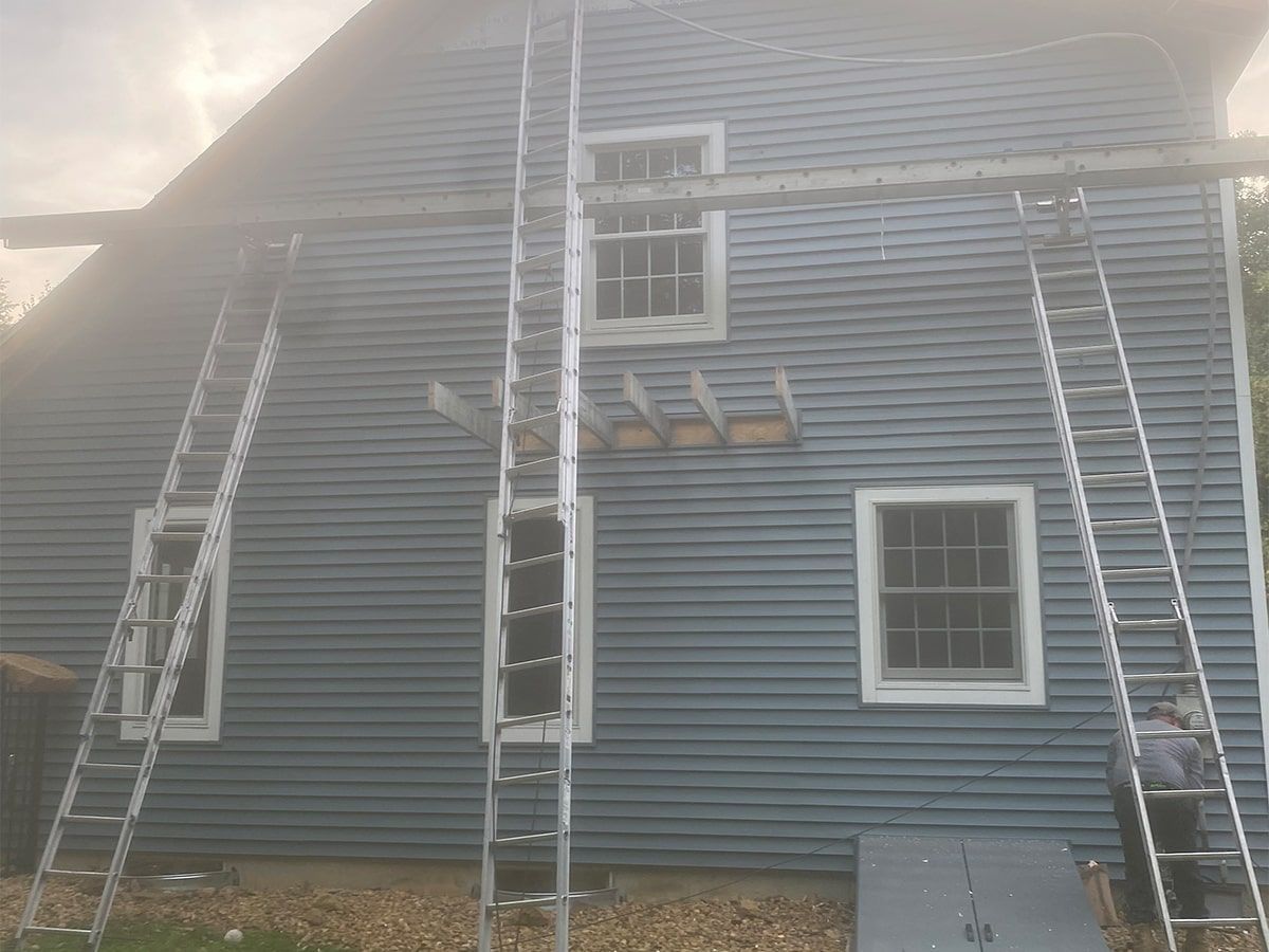 Blue house exterior undergoing repairs with three ladders positioned against the wall and exposed ceiling joists.