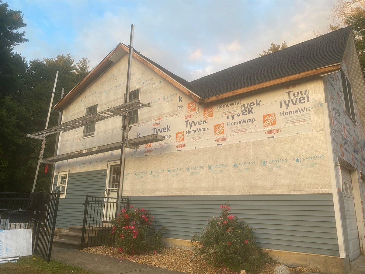 A two-story house under renovation, featuring partial grey siding, exposed building wrap, and wooden scaffolding.