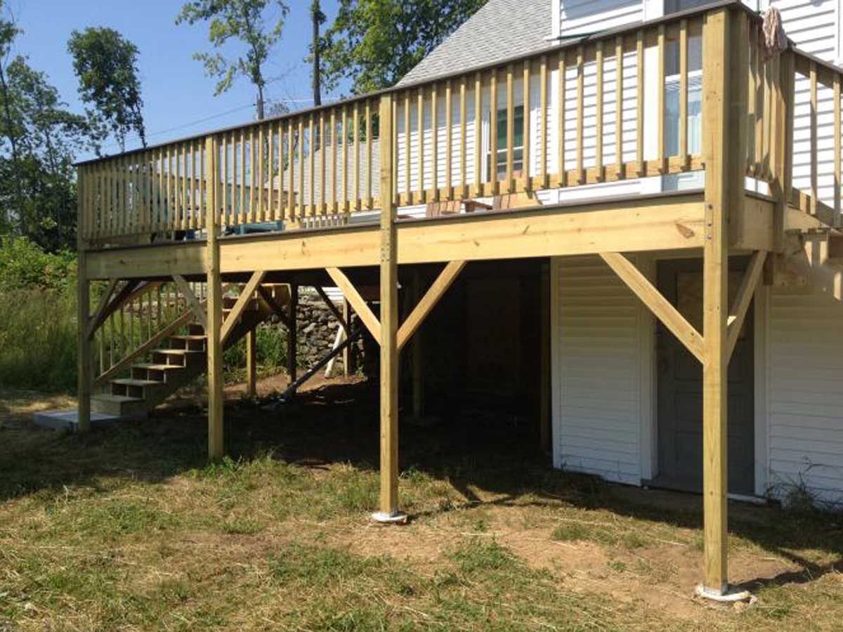 A wooden deck attached to the back of a house, featuring stairs leading down to a grass lawn under a bright blue sky.
