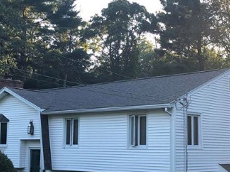 A white, single-story house with a grey shingled roof, set against a background of green trees.