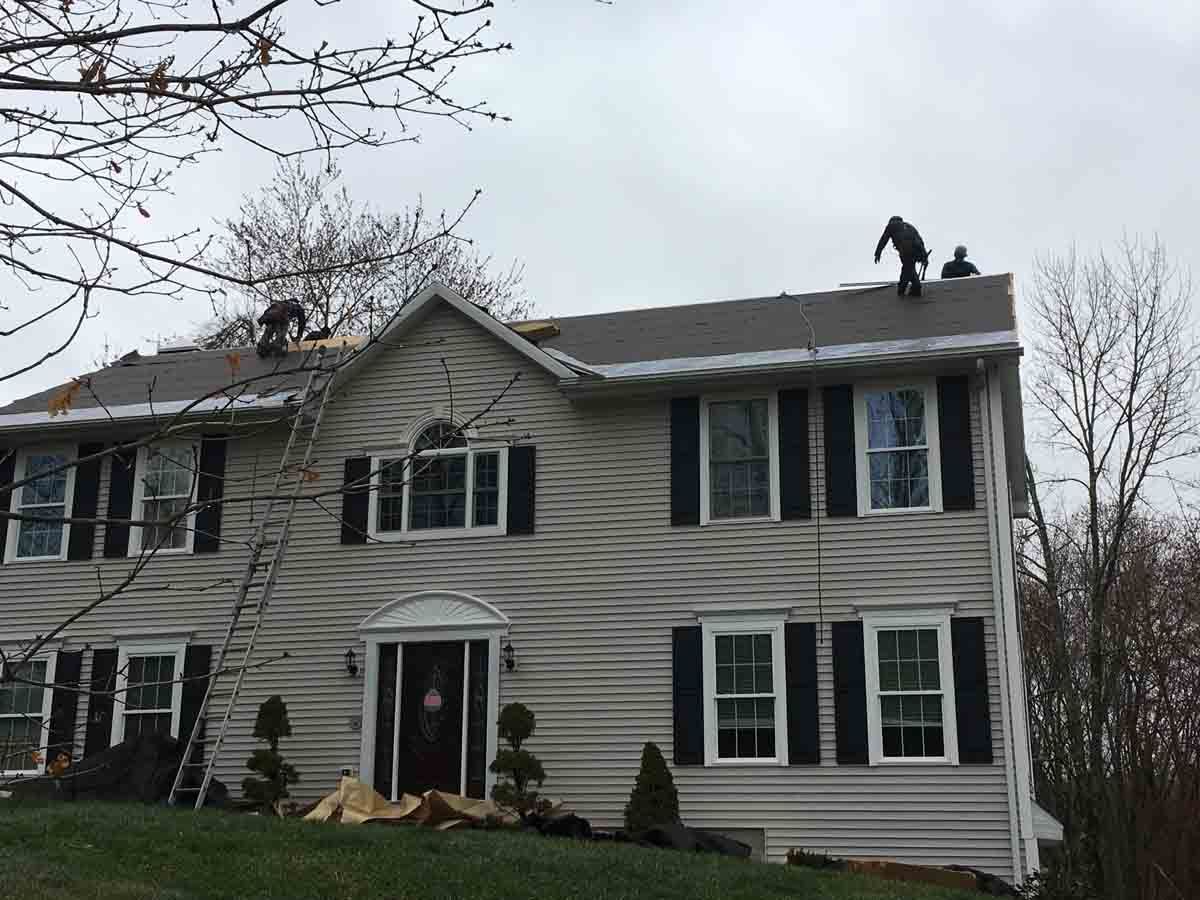 Two workers on the roof of a two-story beige house with black shutters, performing repairs under an overcast sky.