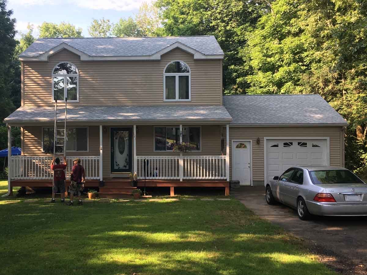 Two workers on a ladder against a beige two-story house with a porch and attached garage, with a silver car parked nearby.