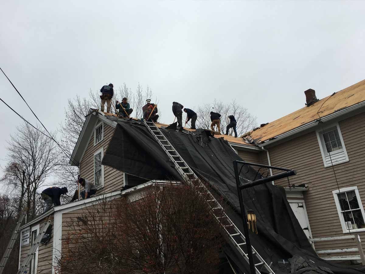 A group of workers on a house roof stripping off shingles, with a black tarp draped down the side of the building.
