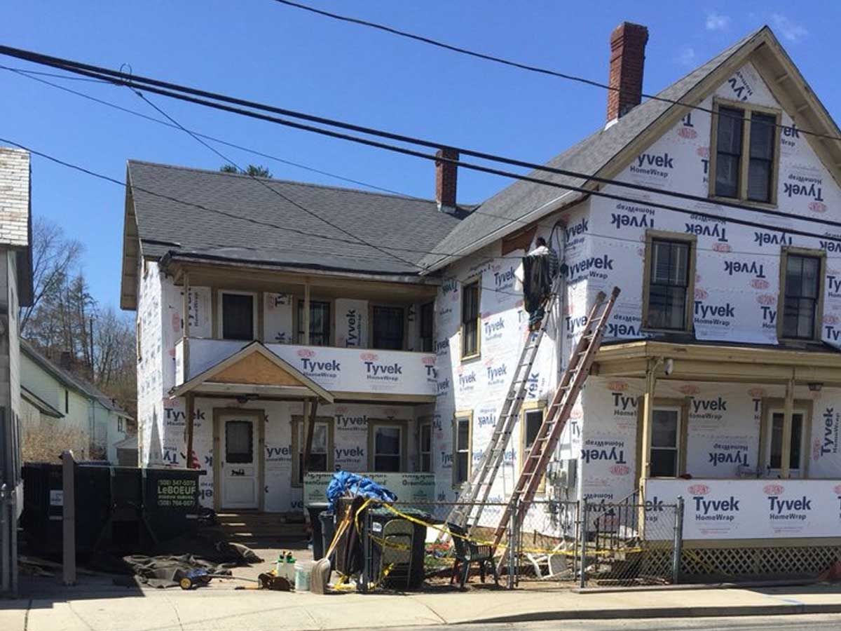 A multi-story house under renovation, covered in white Tyvek weather barrier, with a ladder leaning against the facade.