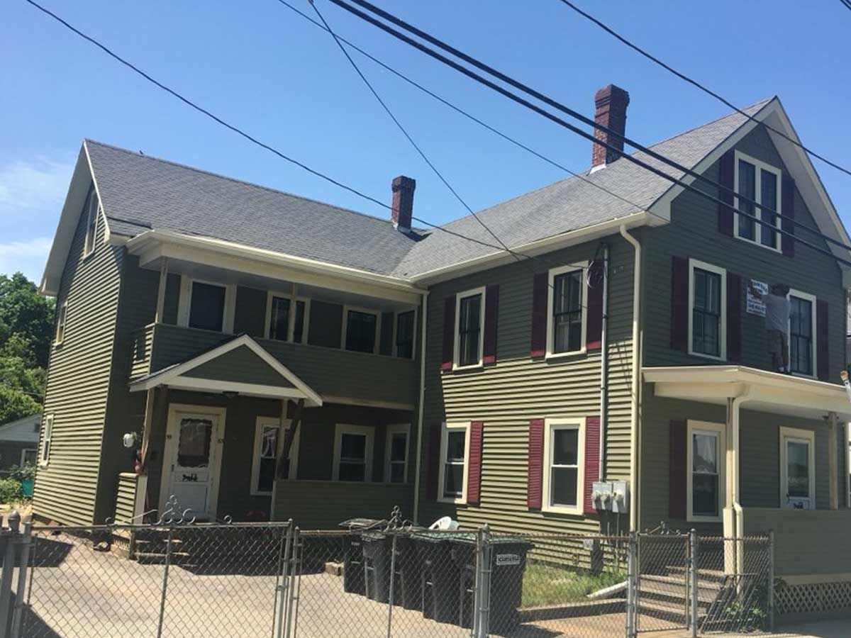 An L-shaped olive green two-story house with maroon shutters and gray shingled roofs, viewed behind a chain-link fence.