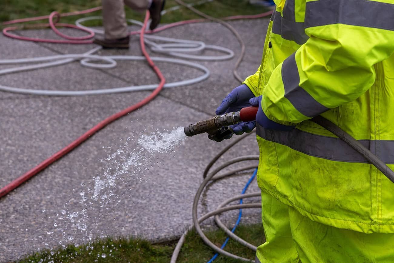 A man in a yellow safety vest is spraying water on a concrete surface with a hose.