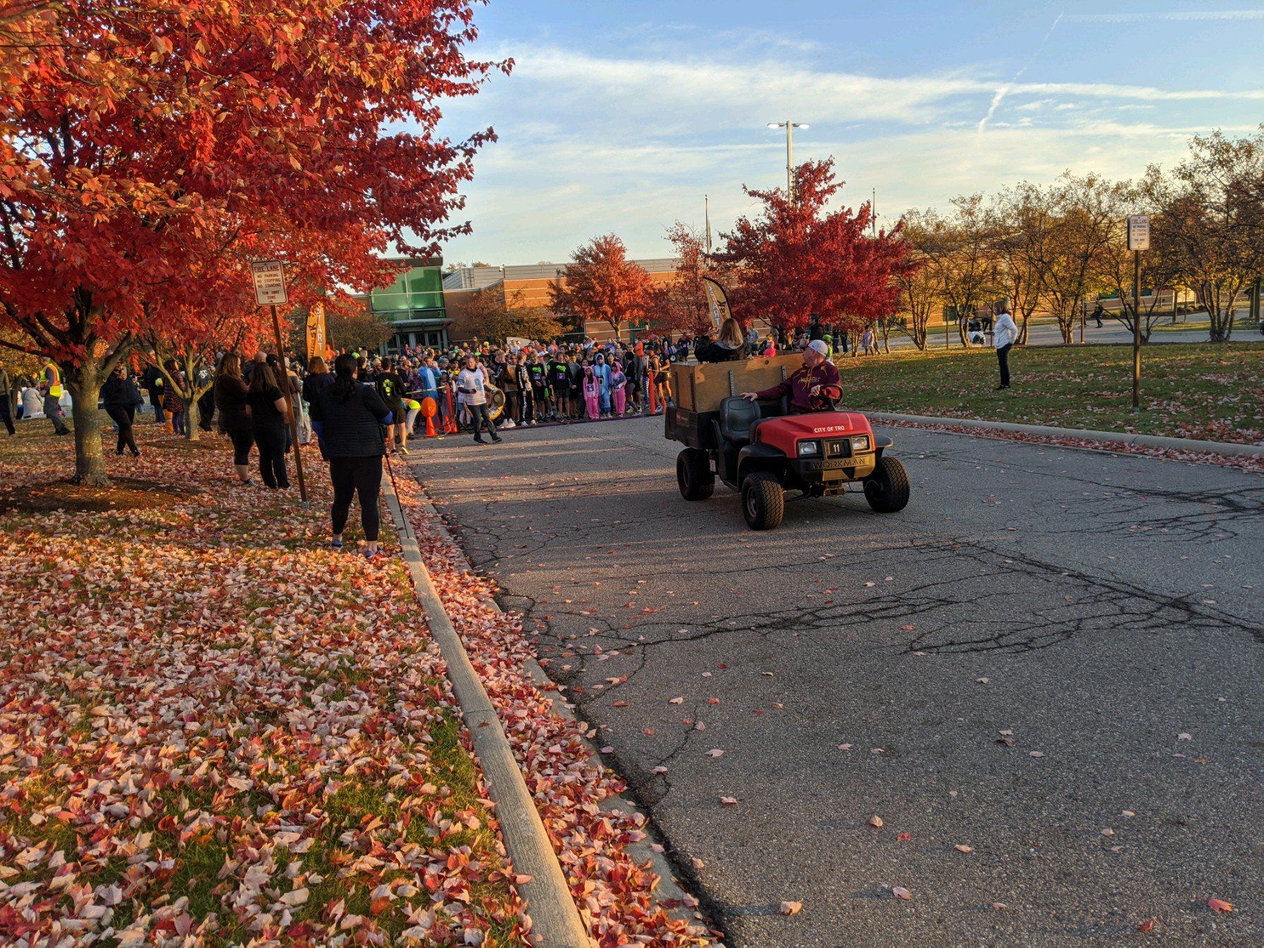 An autumnal scene with red-leafed trees lining a road. A small red tractor pulls a wagon in front of a crowd of people.