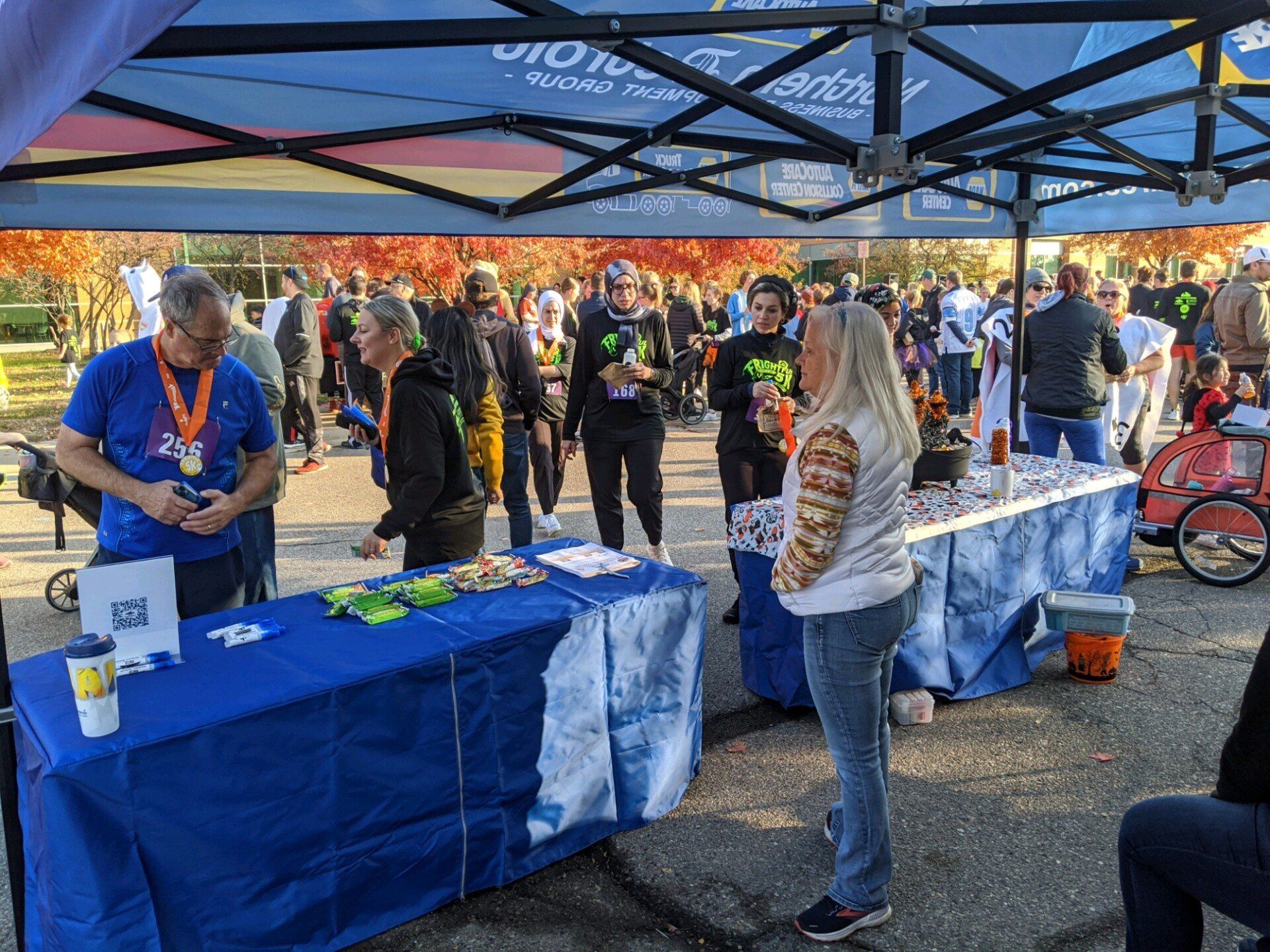 People at a race day tent with tables of food and drinks, under a blue-and-white canopy. Fall foliage and other runners are in the background.