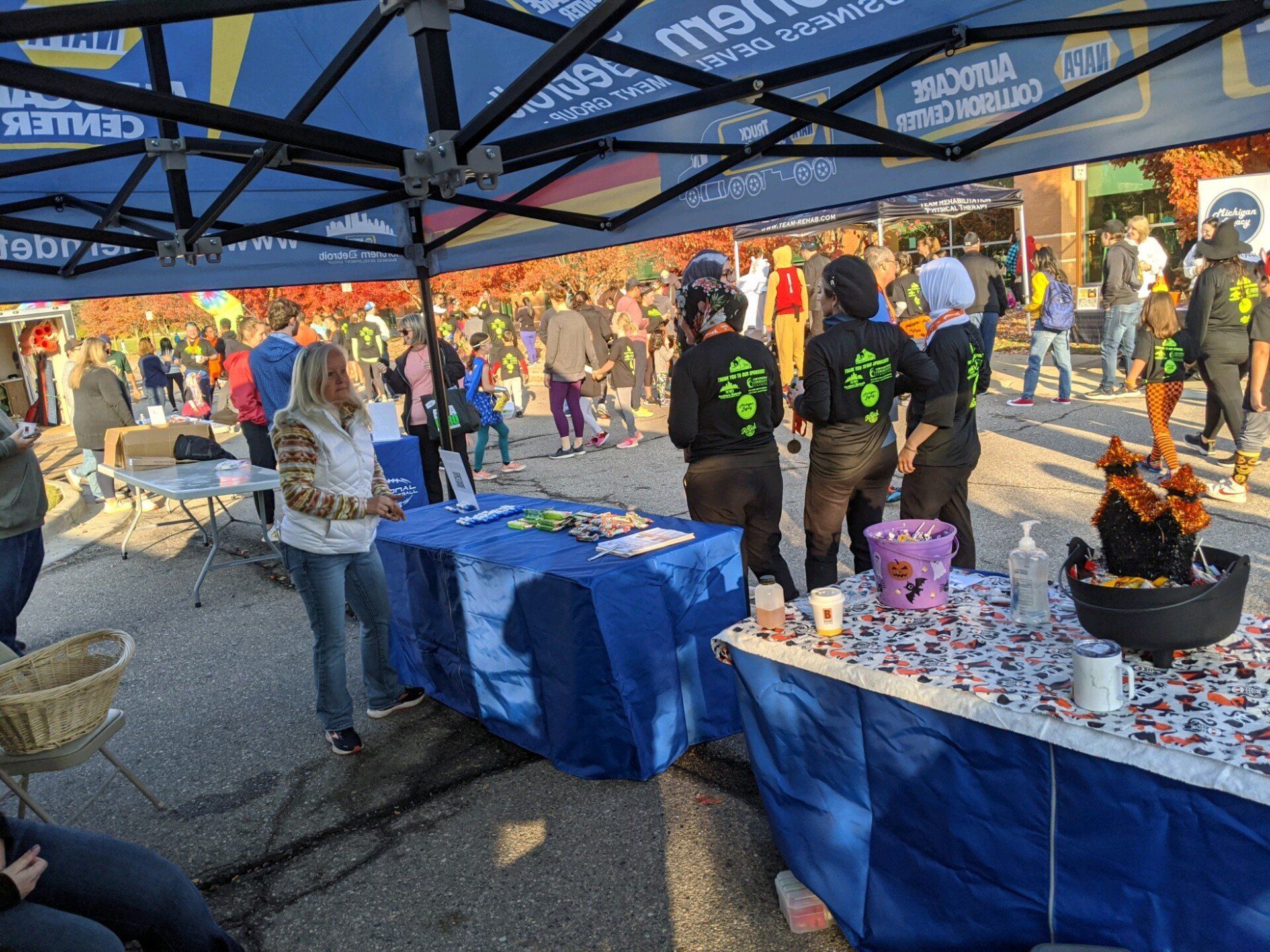 A crowd gathers at an outdoor event. Tables covered in blue cloth and a dark blue canopy provide a booth for event staff, who wear black shirts.