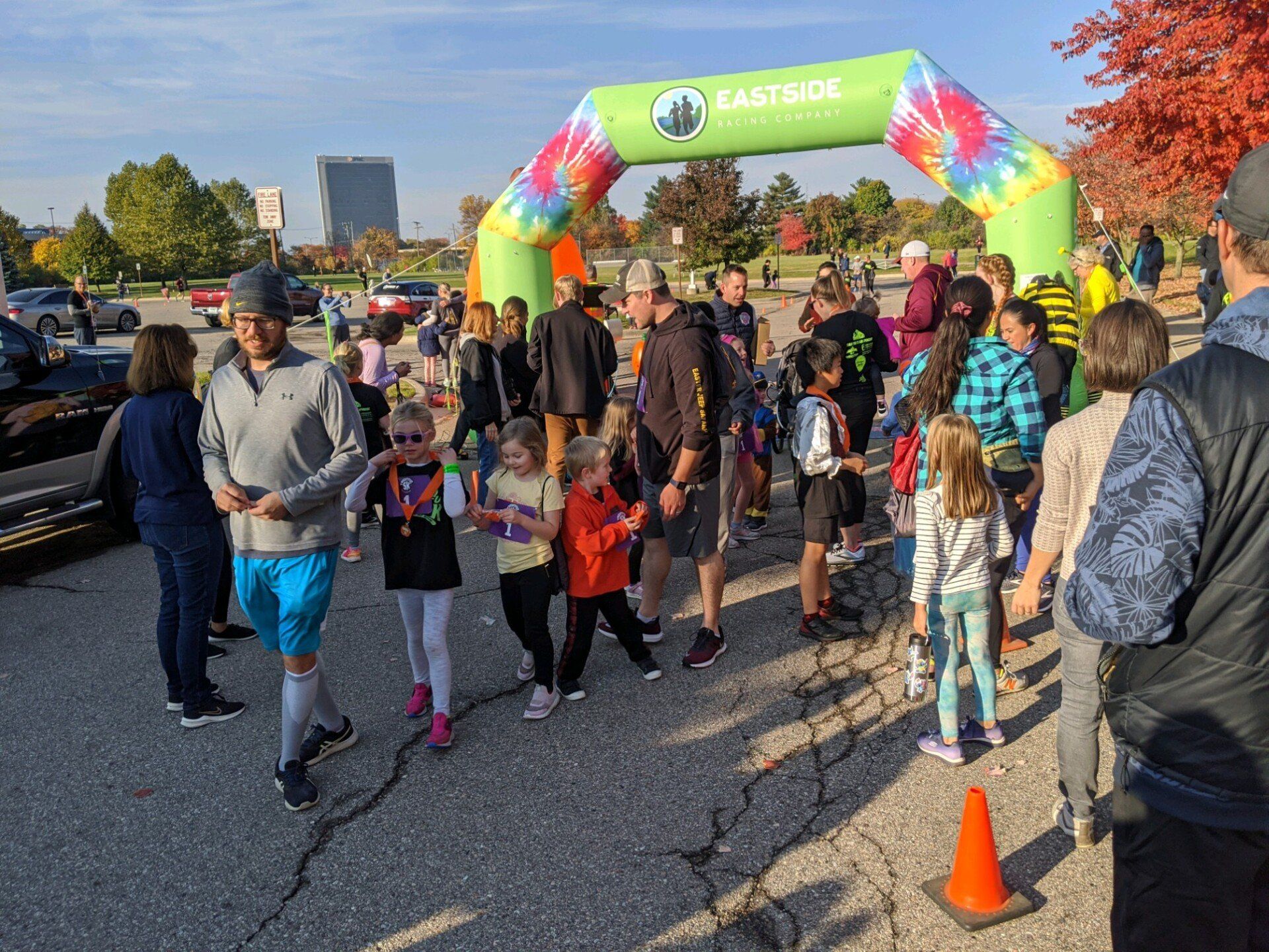 People at a fun run, gathered under a tie-dye archway. Children and adults stand together, some in motion, in an outdoor setting on a sunny day.