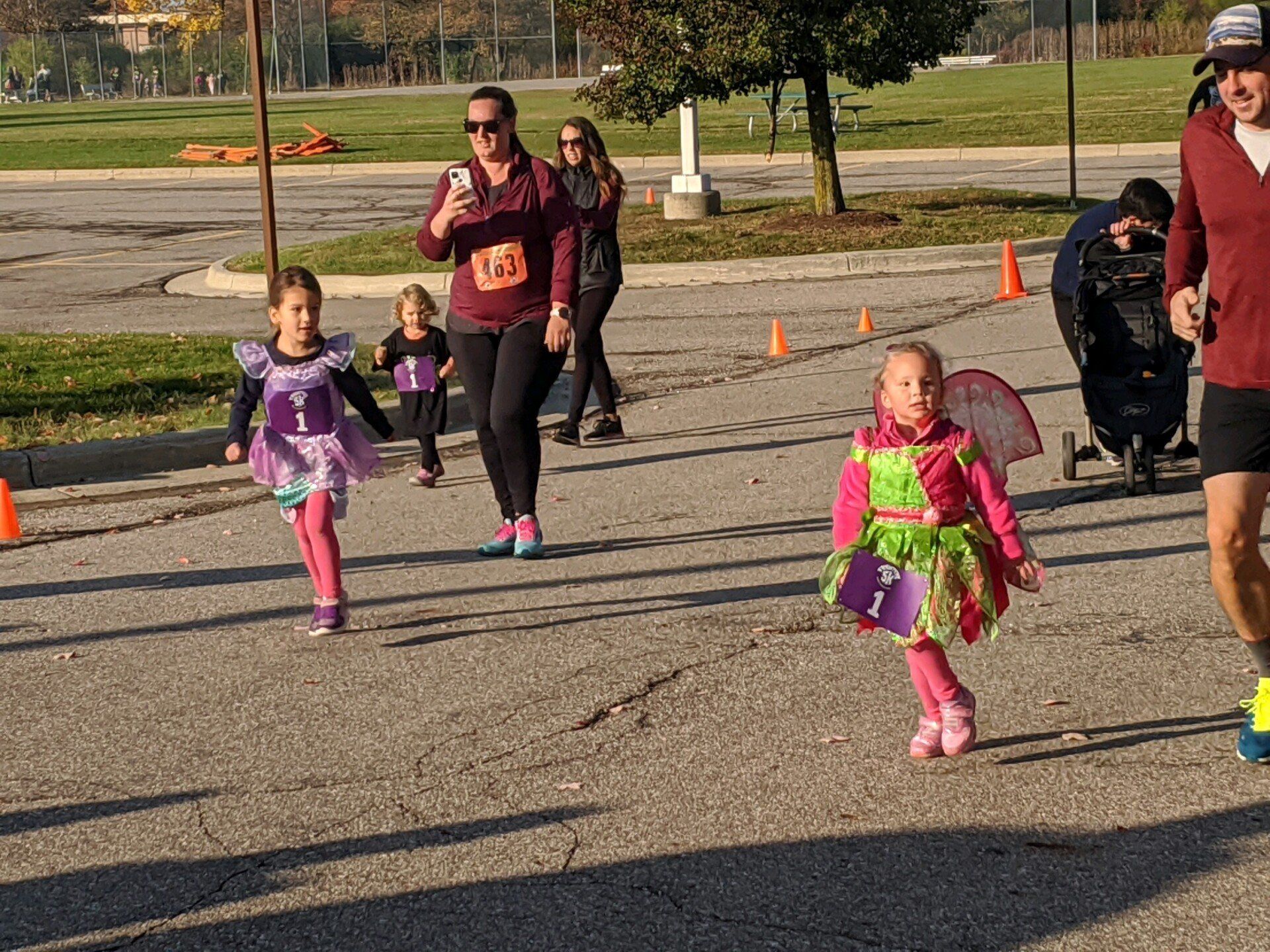 Kids in costumes run a race on asphalt. Two girls wear fairy costumes, while another girl and a woman follow.