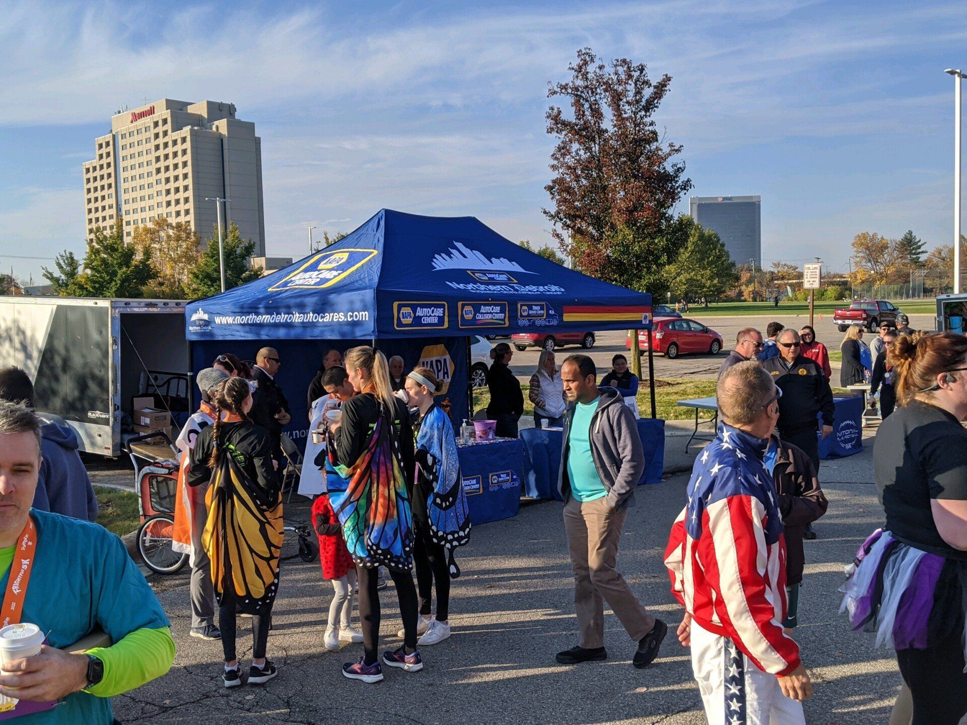 People gather near a blue tent at an outdoor event. Buildings and cars are visible in the background on a sunny day.