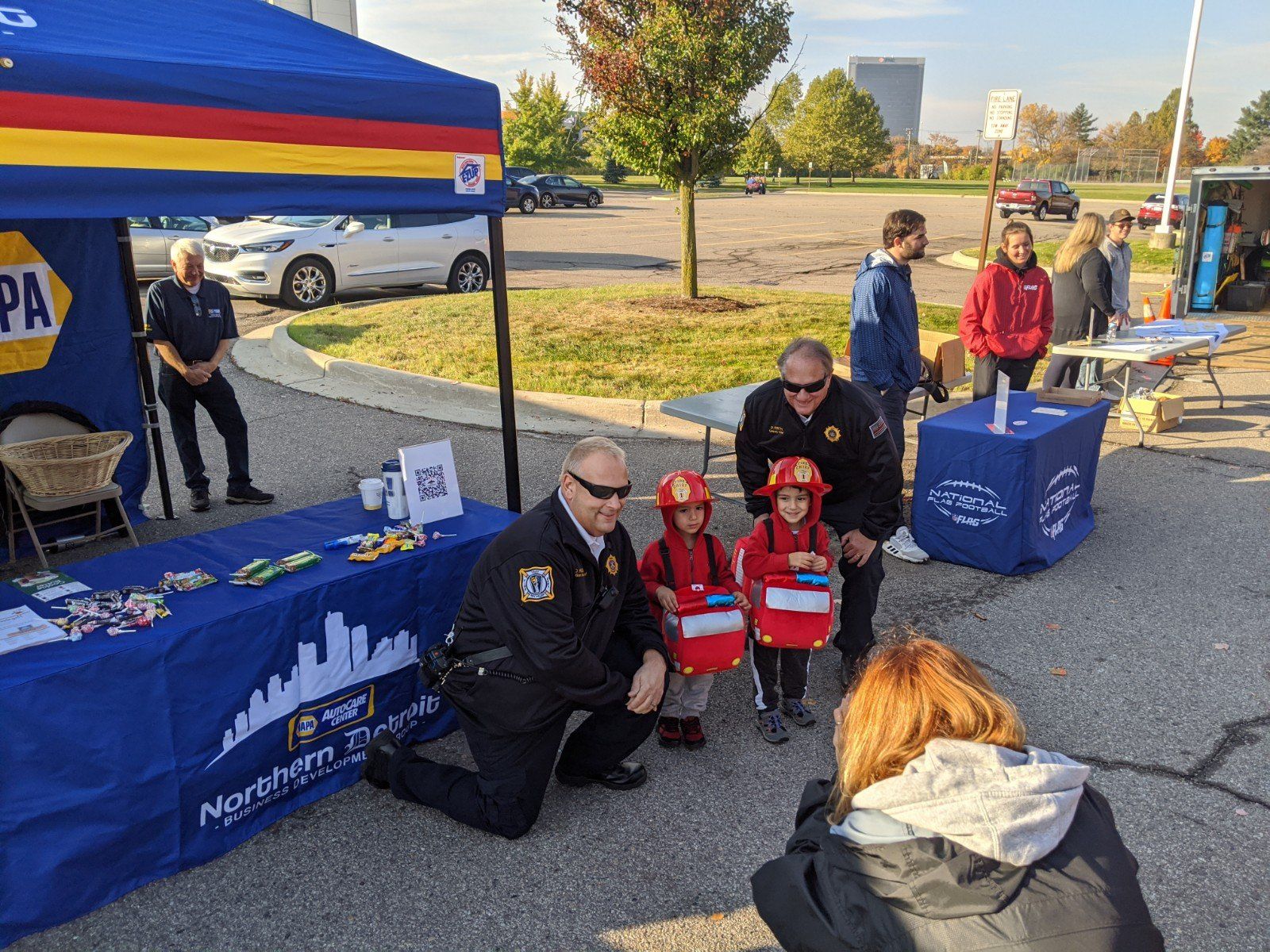 A police officer kneels with two young children in firefighter costumes. The children are posing in front of a NAPA tent.