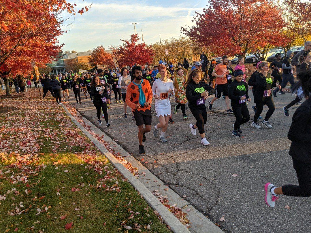 Runners in costumes start a race on an autumn day, surrounded by colorful trees and fallen leaves.