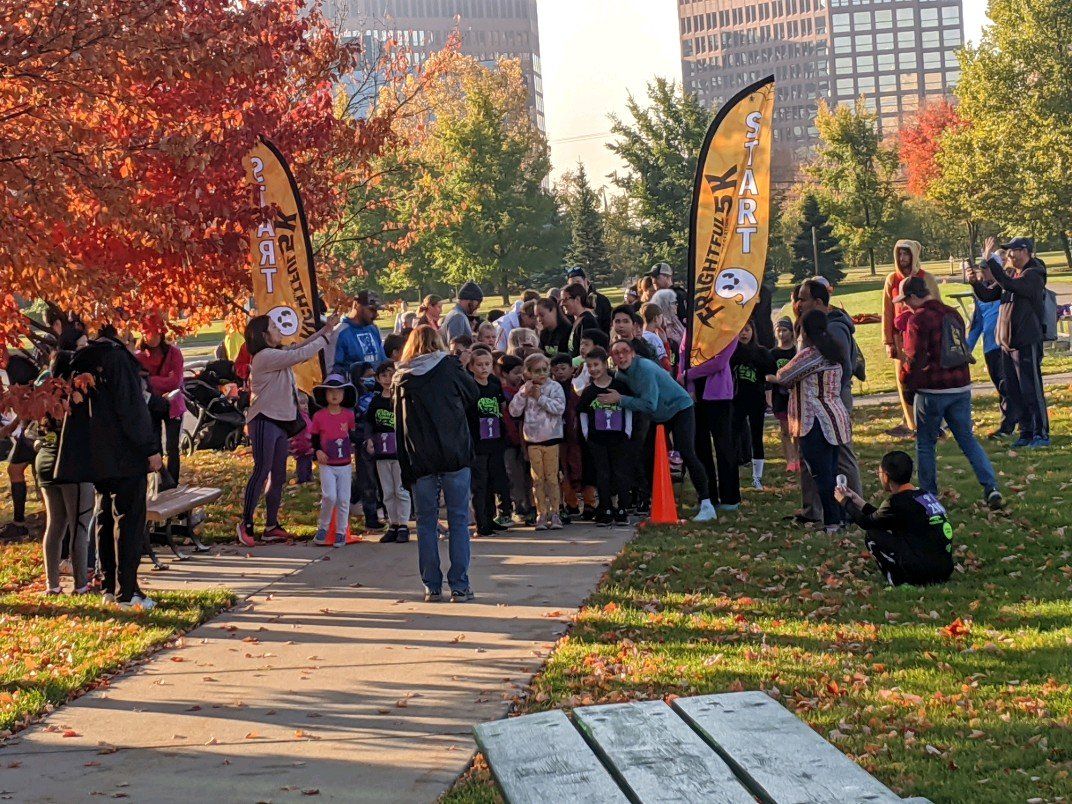 A group of people, including children, gathered at a park for an outdoor event. Fall foliage and tall buildings are in the background.