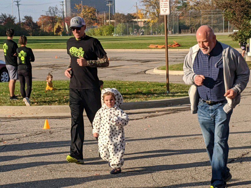 Man in black shirt running with child in Dalmatian costume; older man runs alongside on asphalt, sunny outdoor setting.