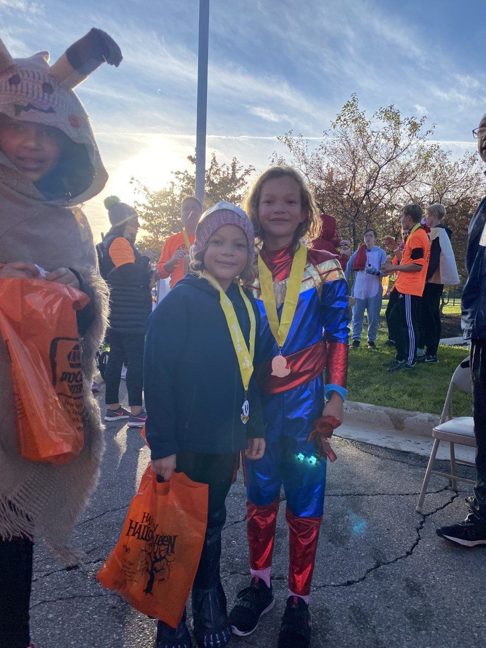 Two children in costumes smiling with medals; one is dressed as Captain Marvel. Outdoor setting with other people visible.