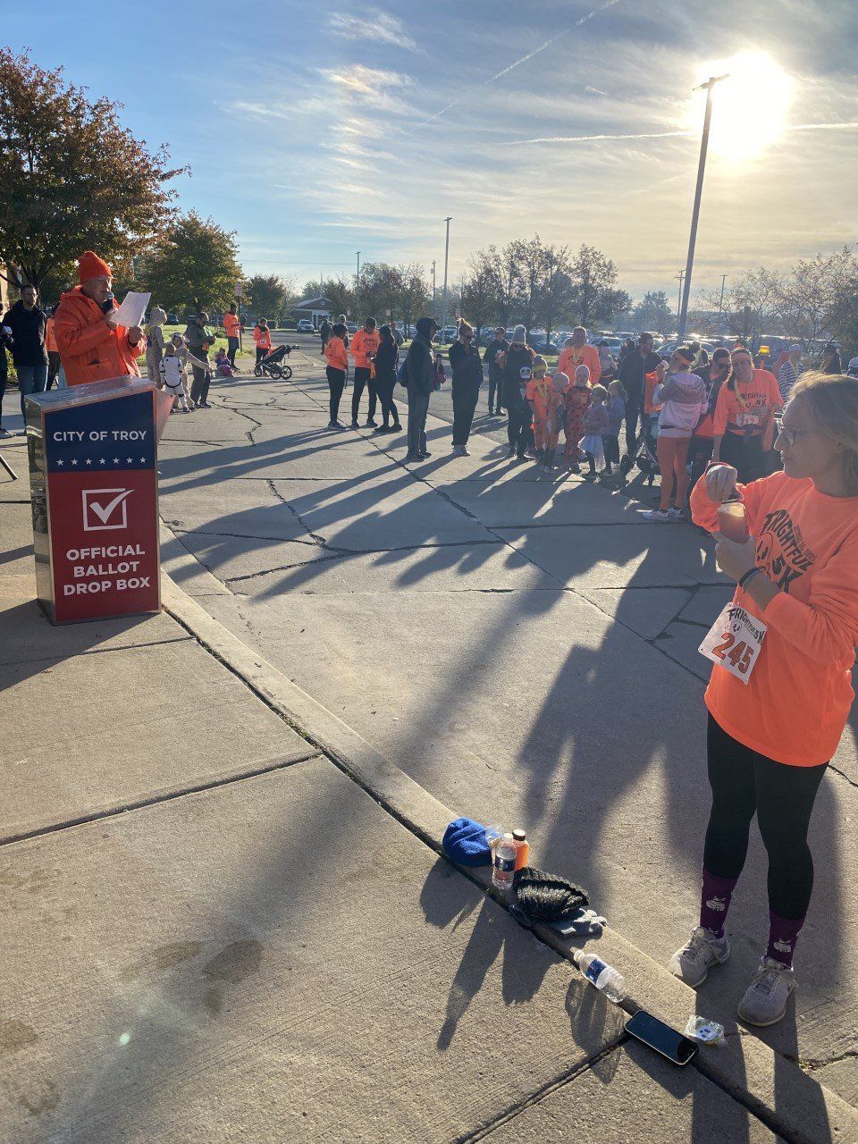 A group of people in orange shirts gathers at an outdoor event with a speaker at a podium. The sun shines brightly.
