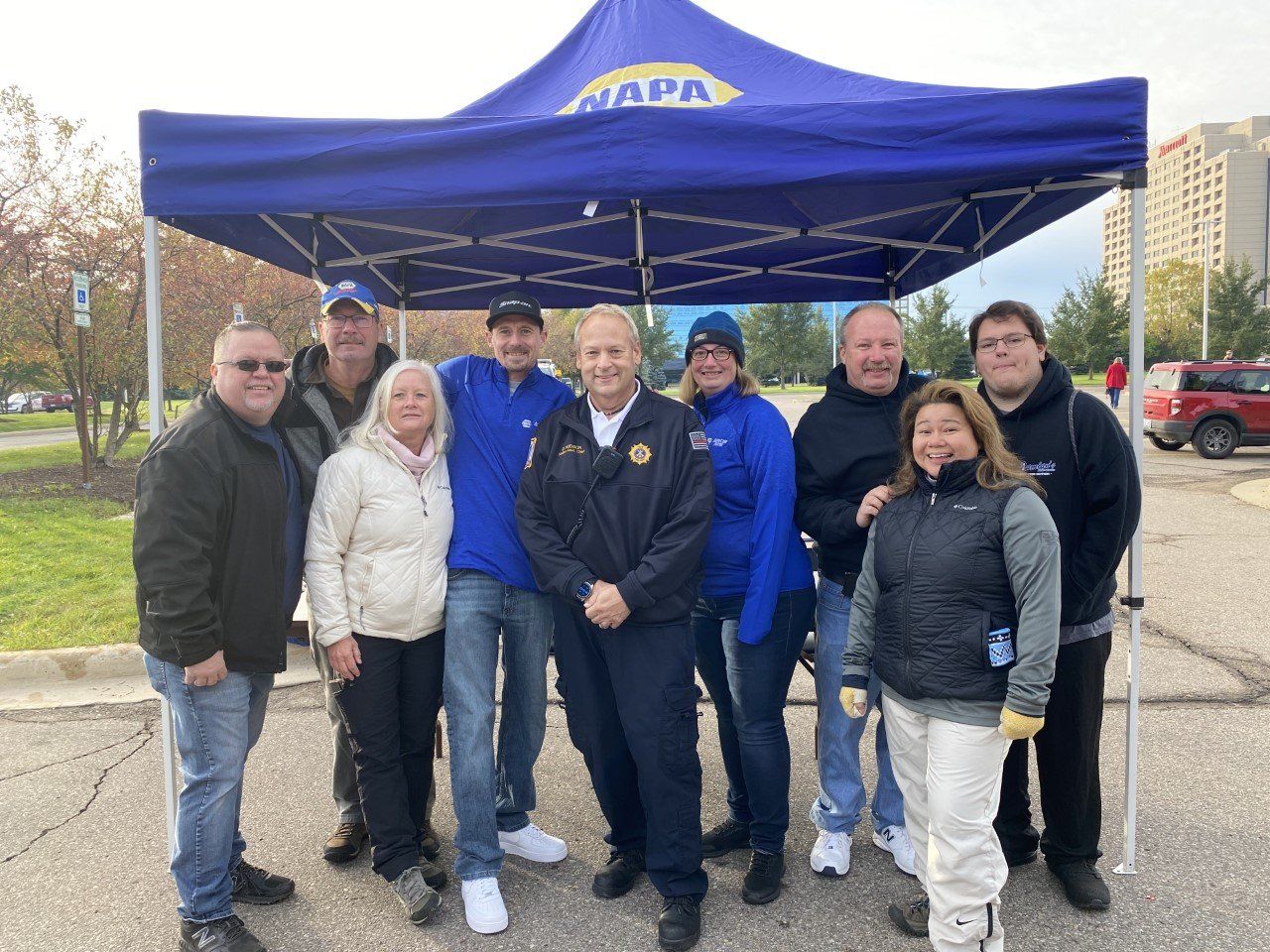 A group of nine people pose under a blue NAPA tent outside. The group includes men and women in casual wear.