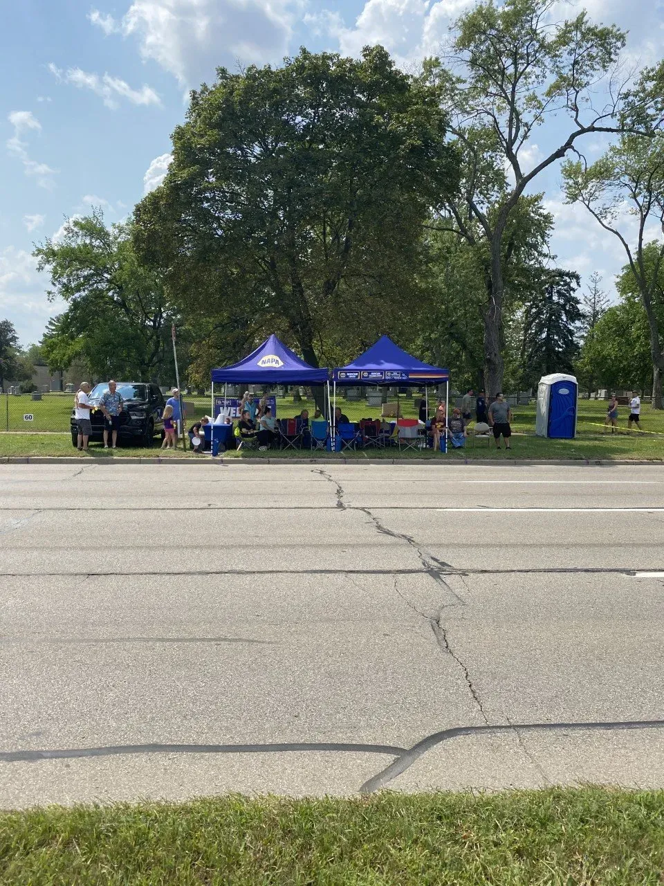 Two blue tents set up roadside, with people gathered. A portable toilet stands nearby.