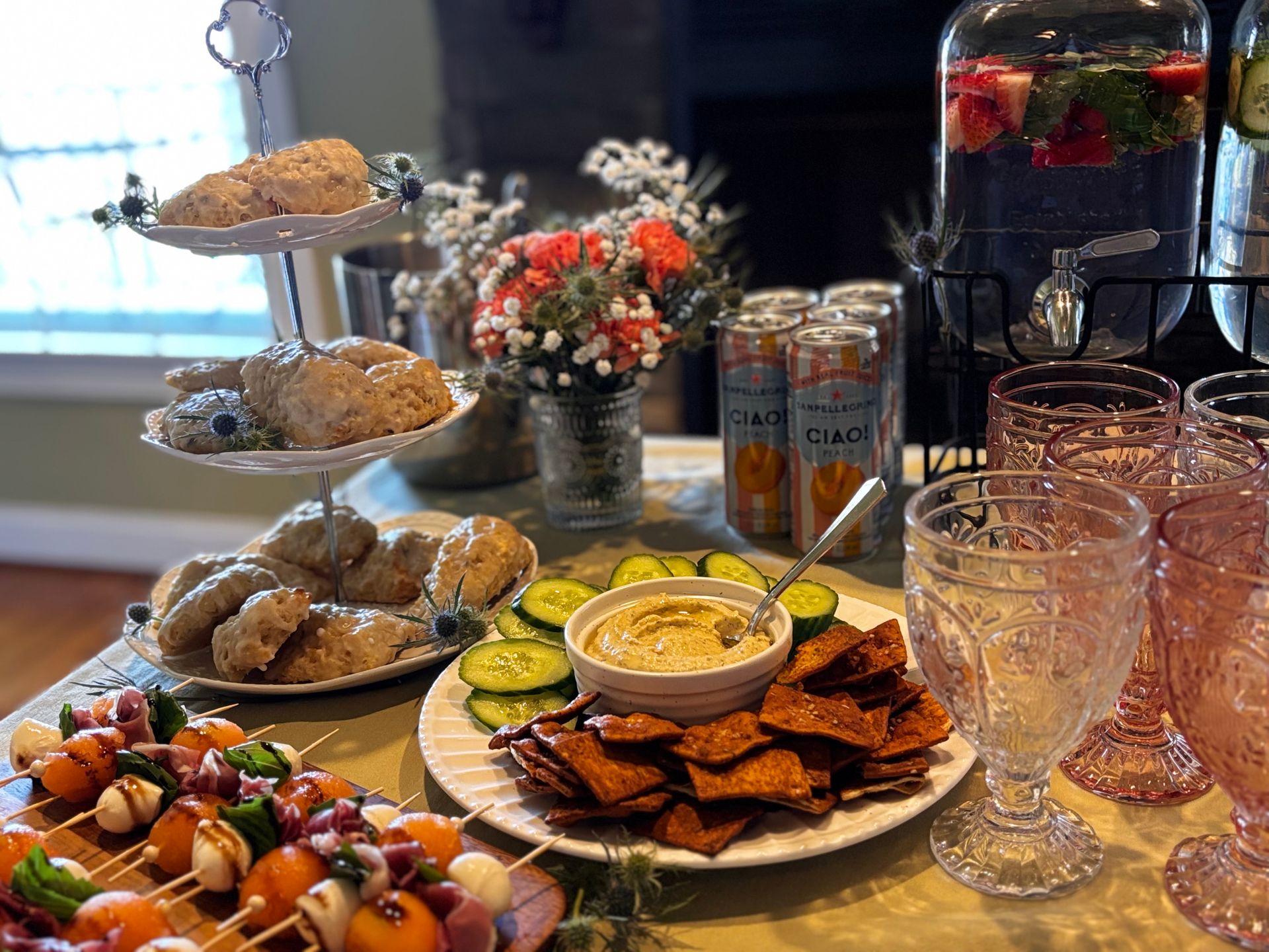 A spread for a tea party featuring scones on a tiered tray, skewers, hummus with cucumbers, and drinks in a dispenser.