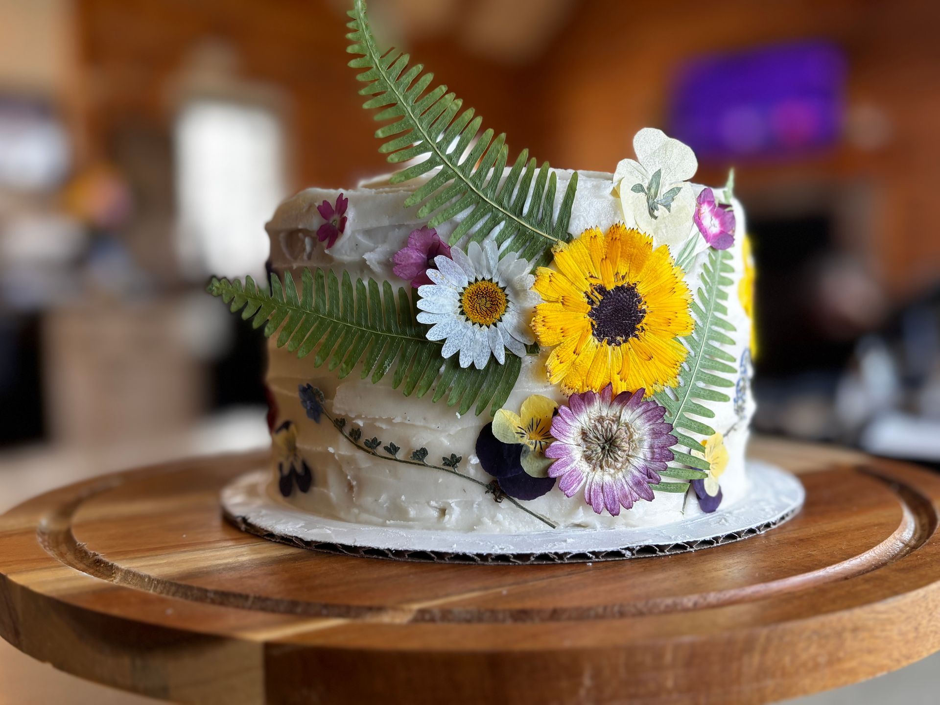 A white frosted cake decorated with pressed flowers and ferns on a wooden cake stand.