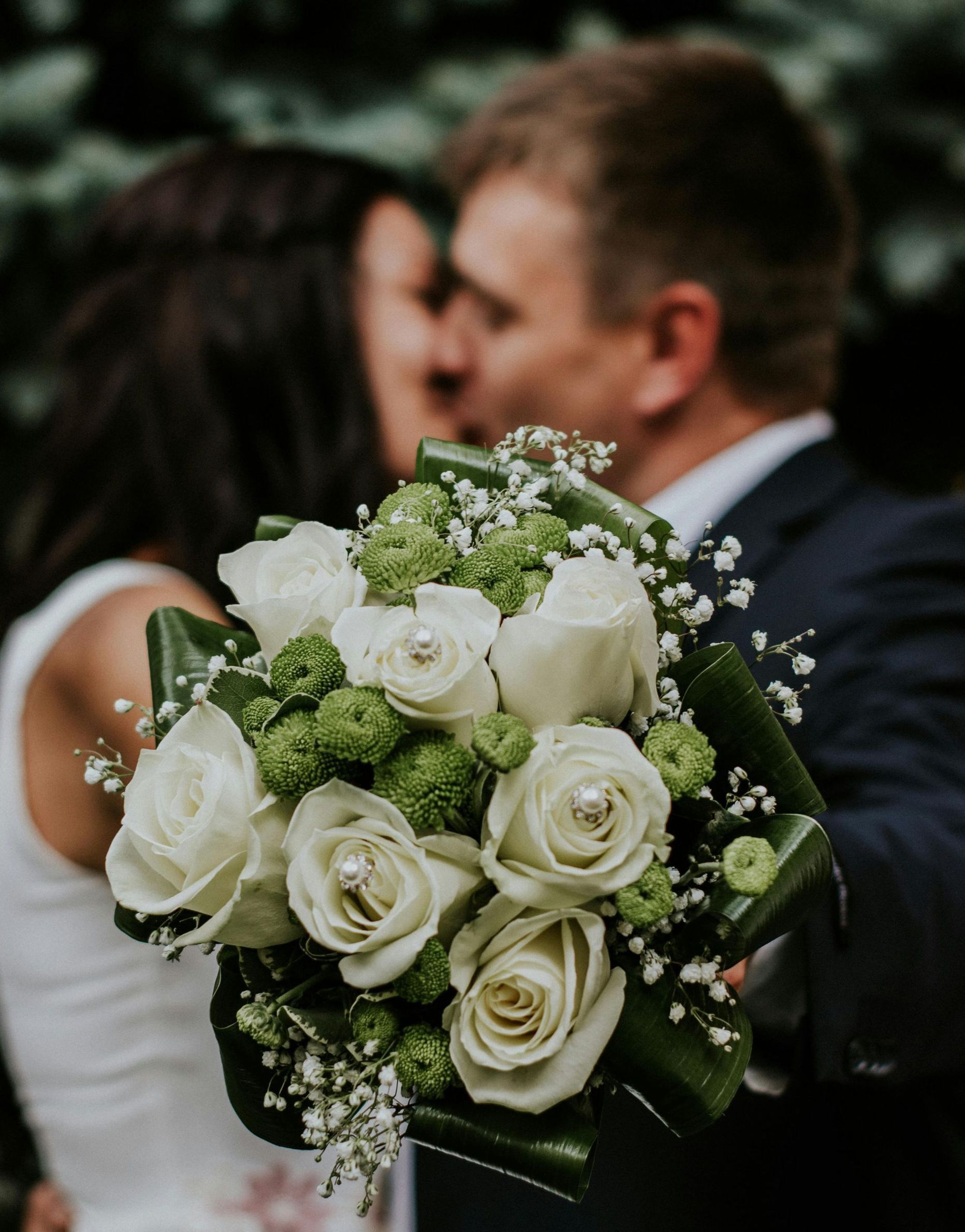 A bride and groom kissing while the bride holds a bouquet of white roses and greenery.
