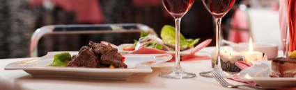 Two glasses of red wine on a table set for dinner; food and a candle in background.