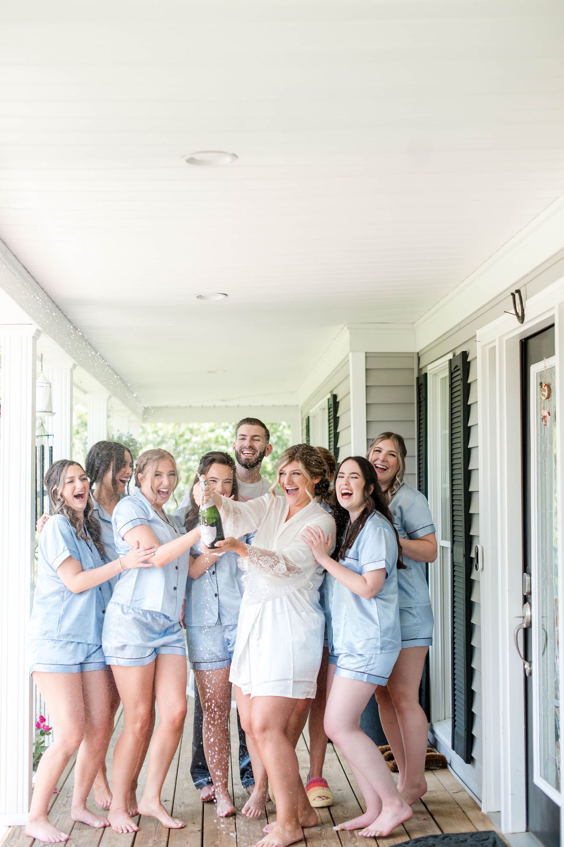 Bride and bridesmaids celebrating, popping champagne on a porch. Wearing pajamas, laughing.