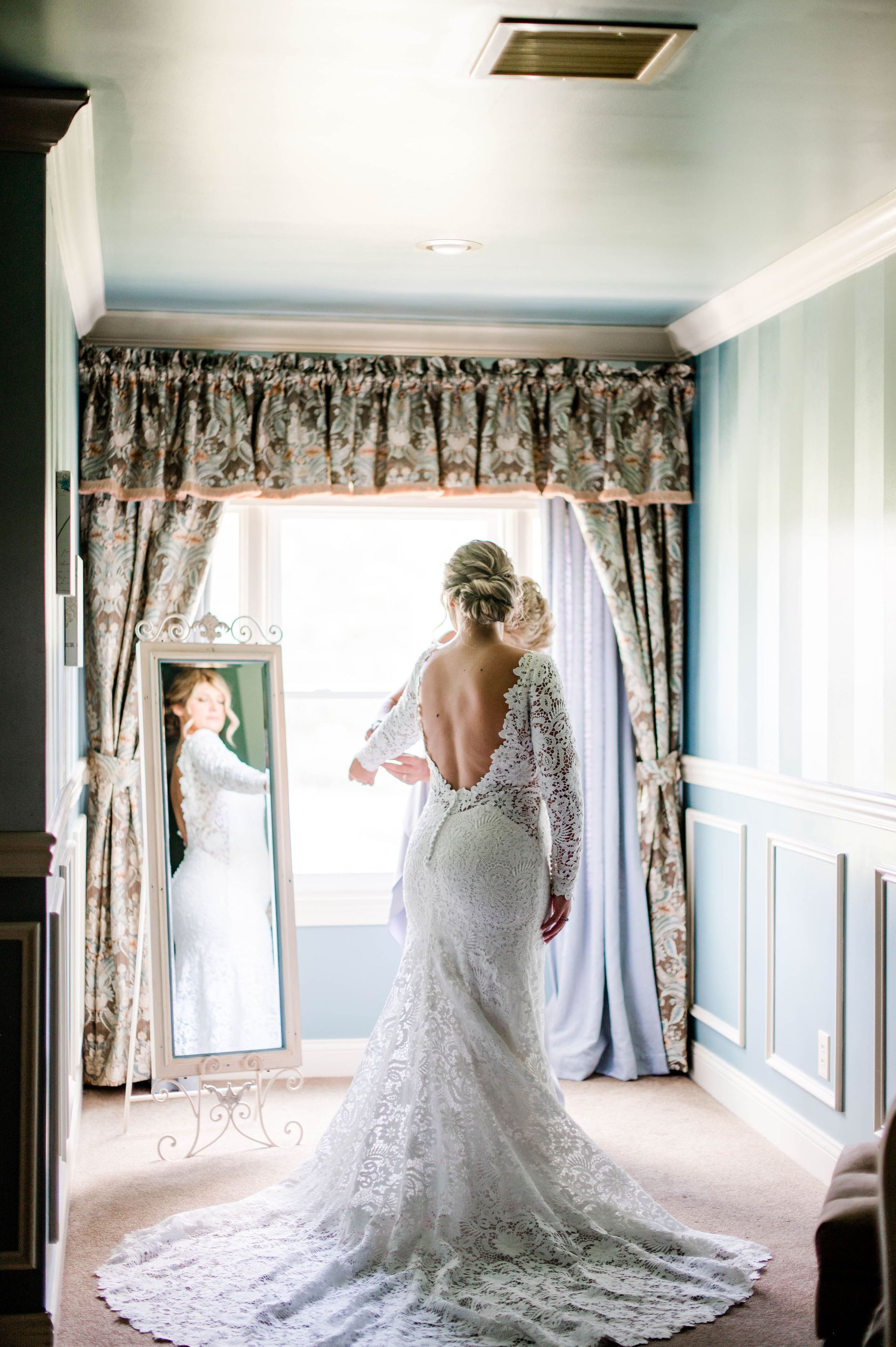 A woman in a wedding dress is standing in front of a mirror in a room.