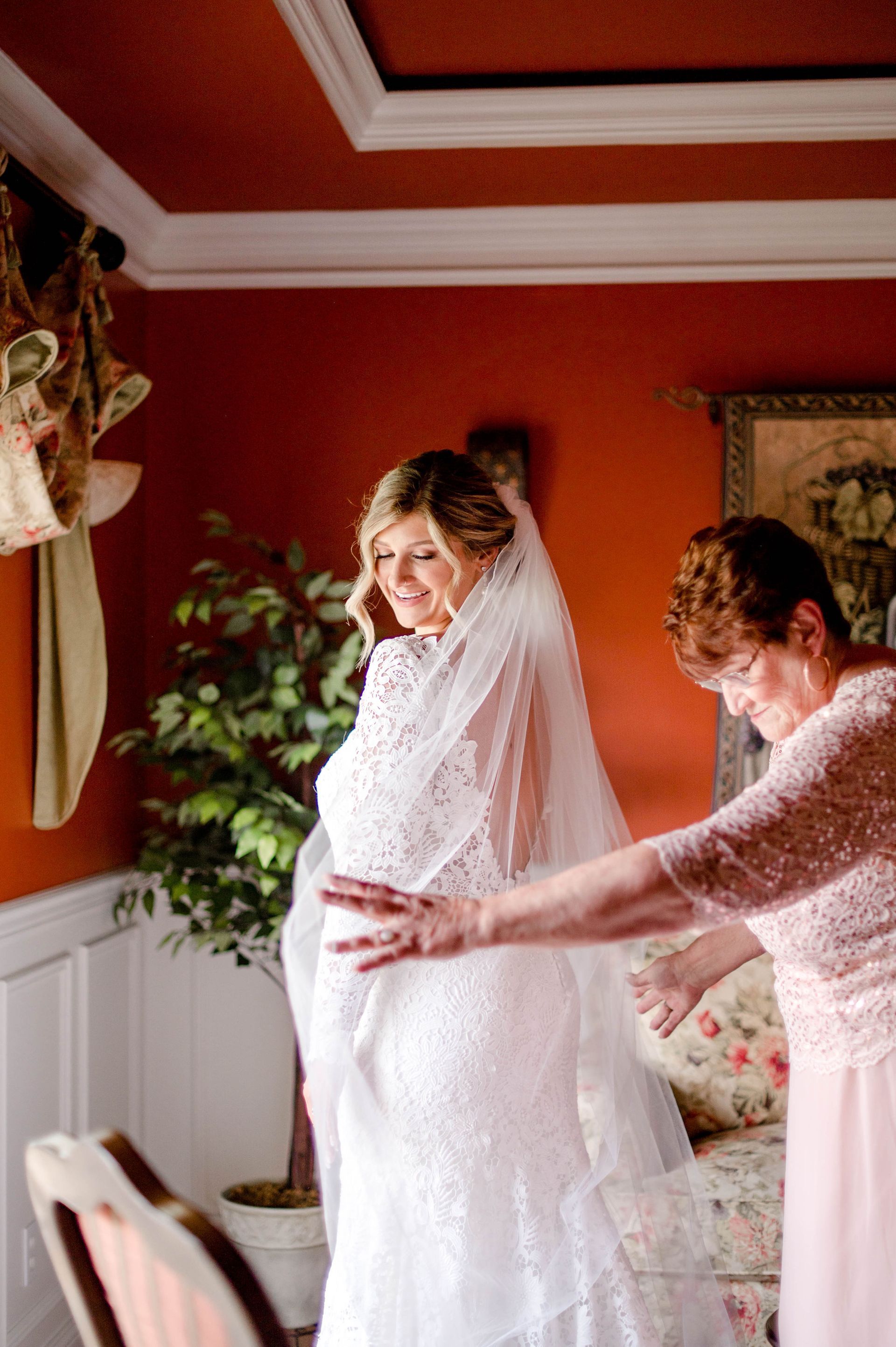 Bride in a white lace dress with veil, helped by a person in a pink dress, in a room with red walls.