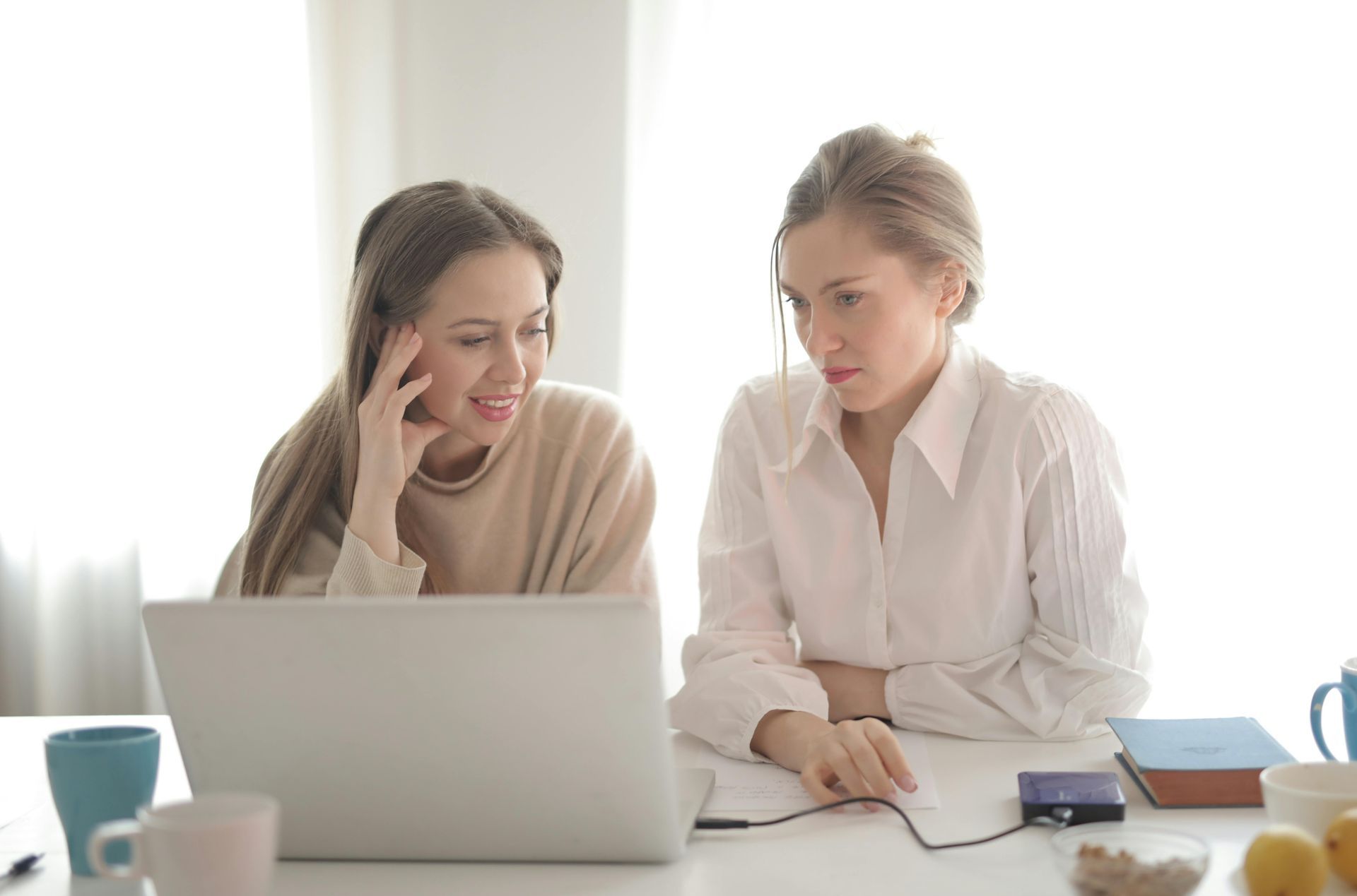Two women looking at a laptop, one smiling, one looking intently. White table, cups, and light background.