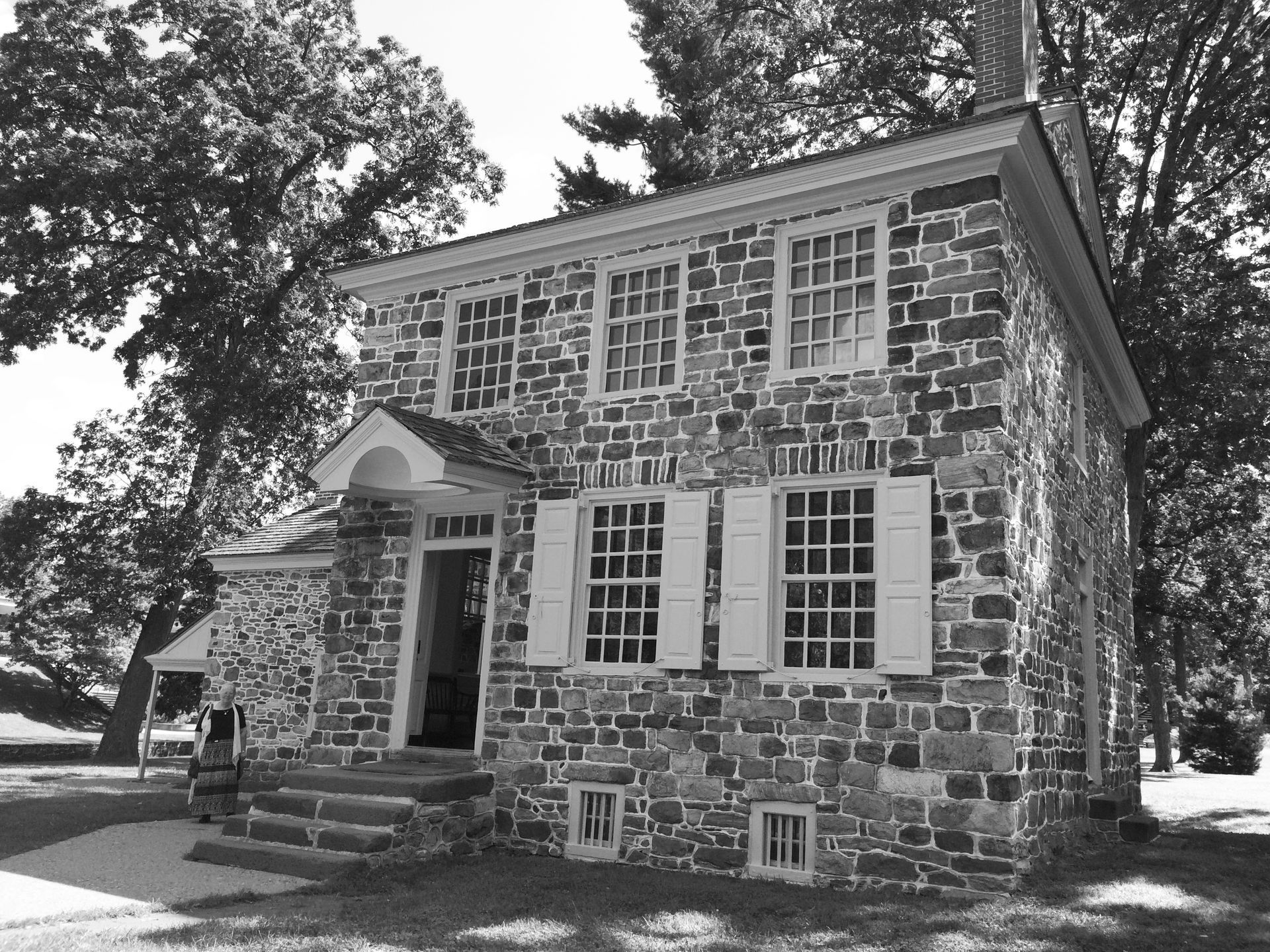 Stone house with multi-pane windows and shutters, set in a grassy yard, trees in the background.