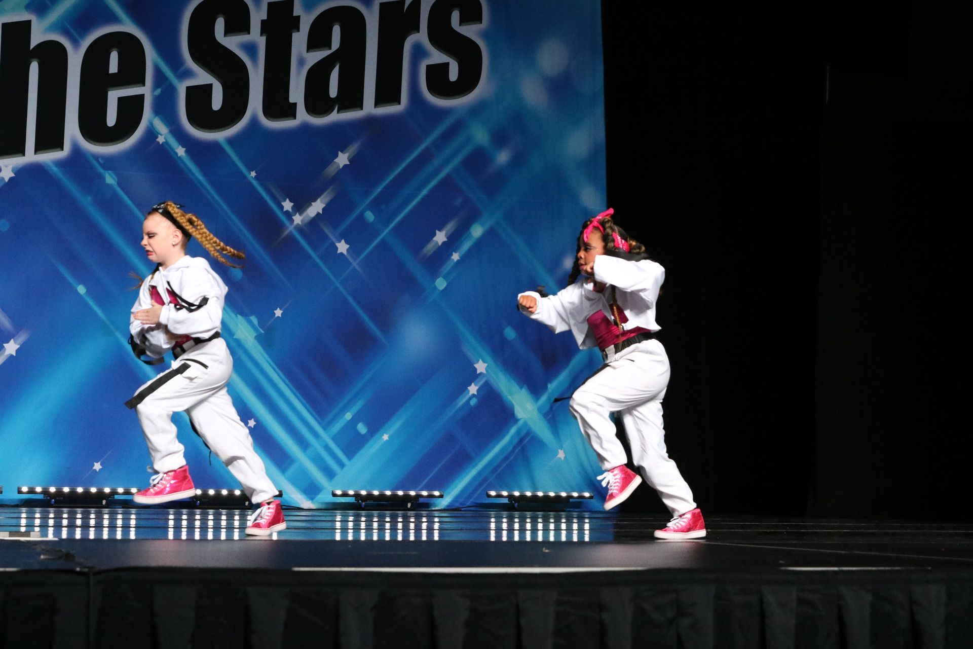 Two dancers in white tracksuits perform on stage with a blue backdrop.