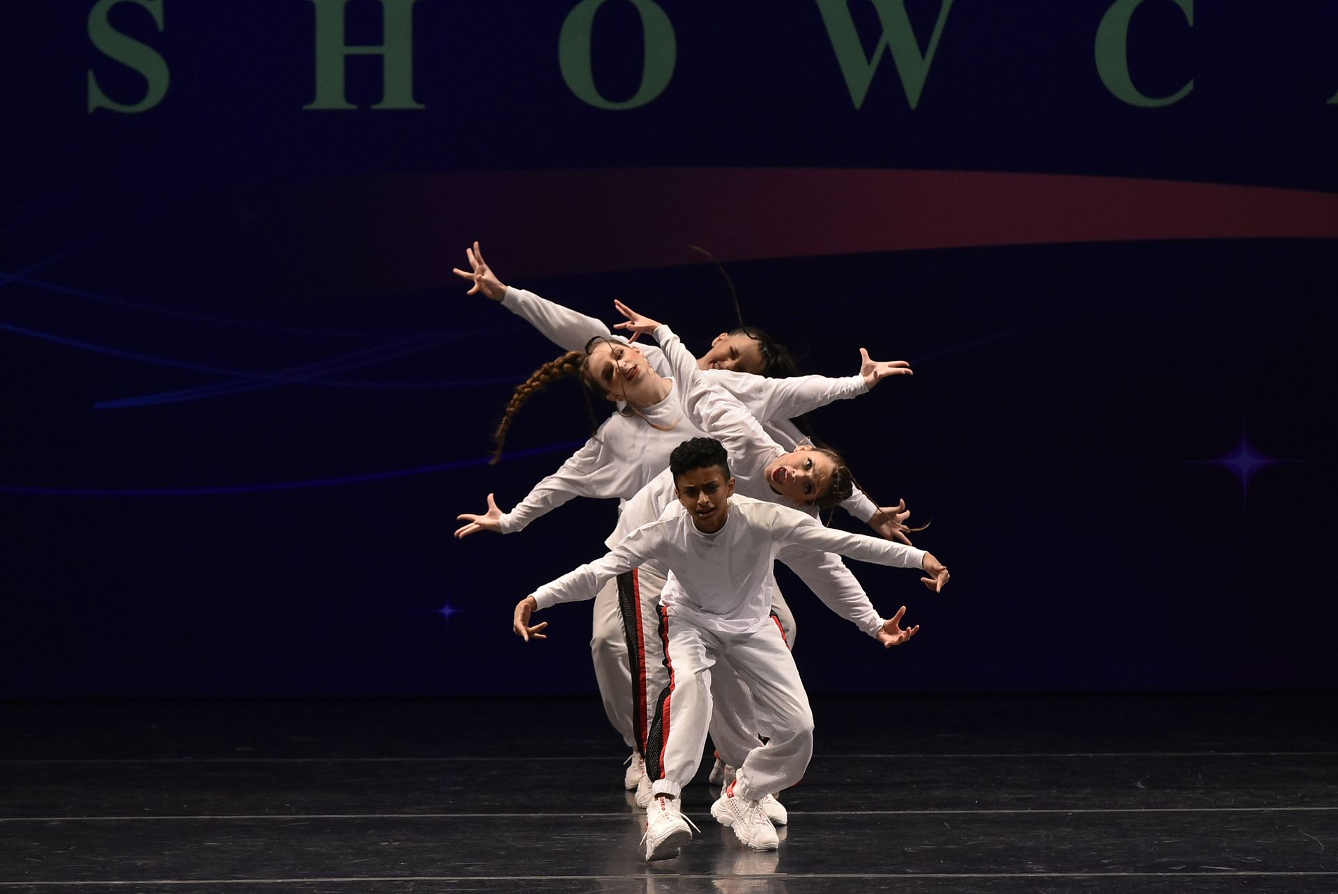 Dancers in white outfits performing a synchronized routine on a stage with a dark background.
