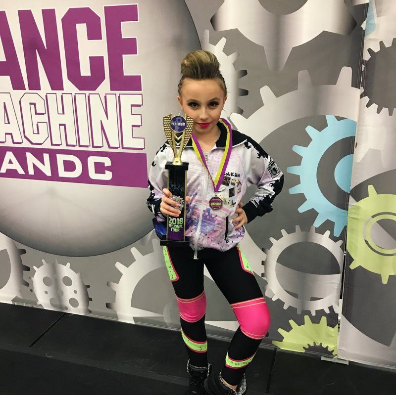 A girl holding a trophy in front of a sign that says dance machine andc