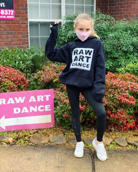 A young girl wearing a mask is standing in front of a raw art dance sign.