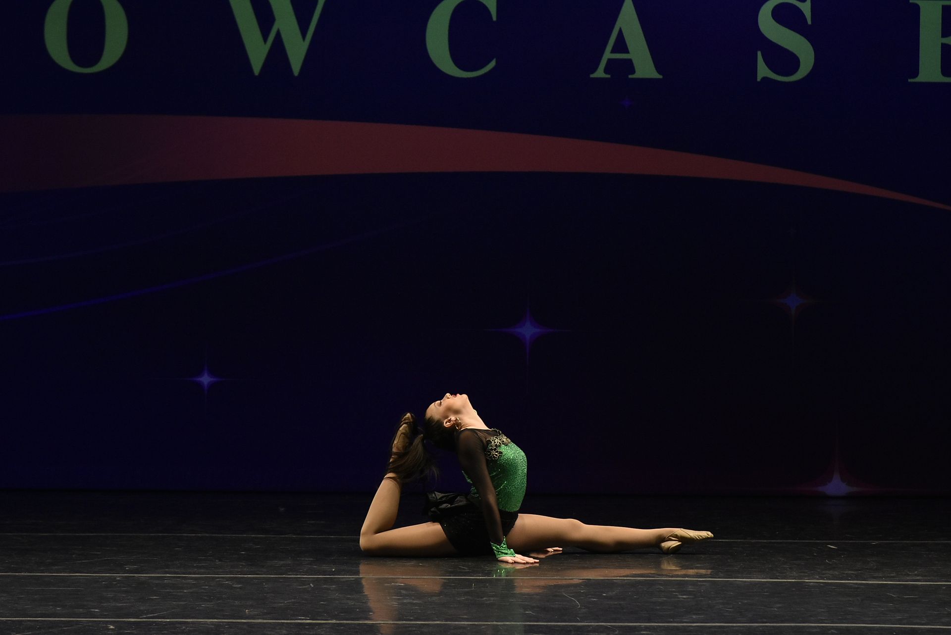 A dancer in a green and black costume arches backward on a stage, with arms extended and head tilted up. 