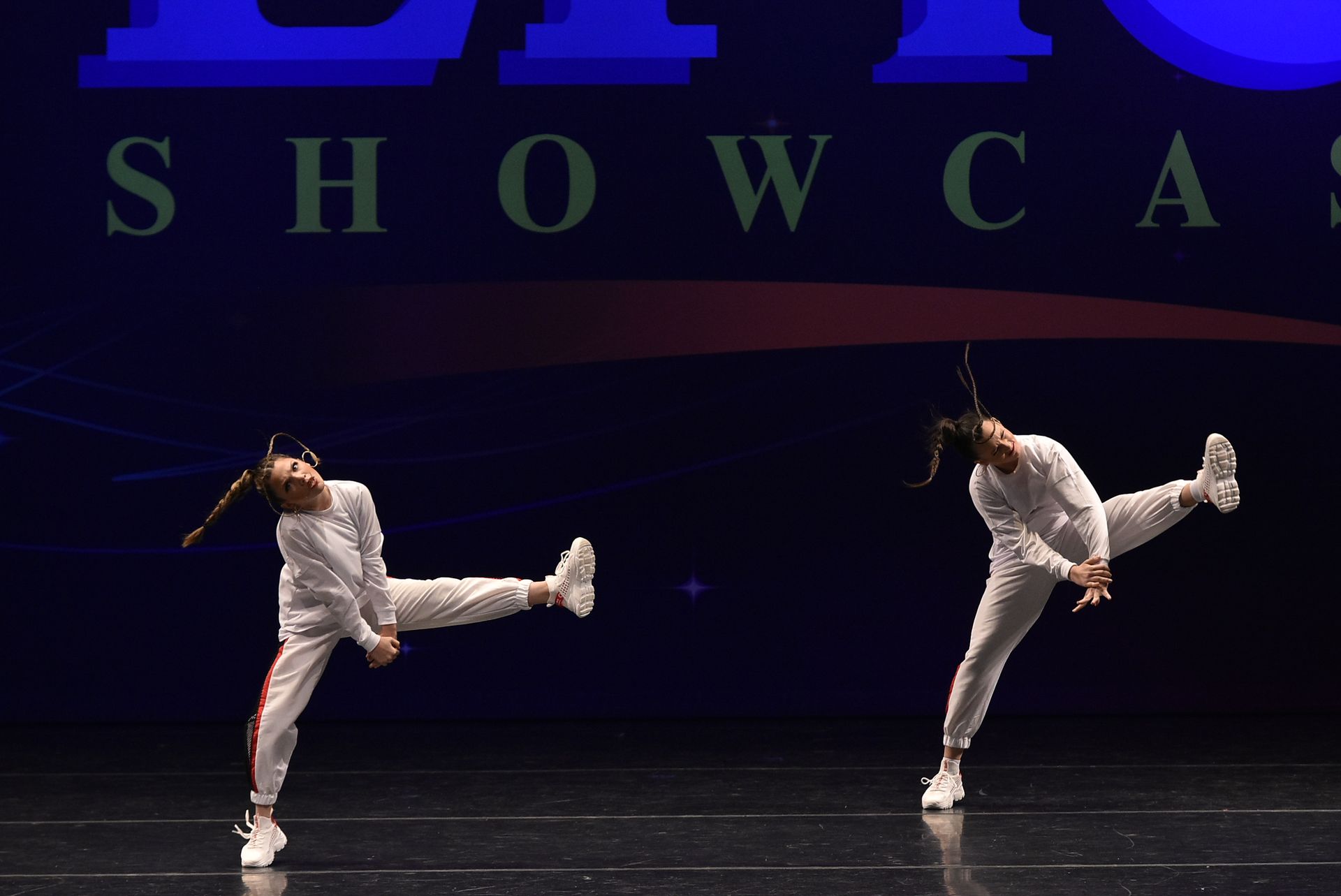 Two dancers in white tracksuits kick their legs up, performing on stage with a 