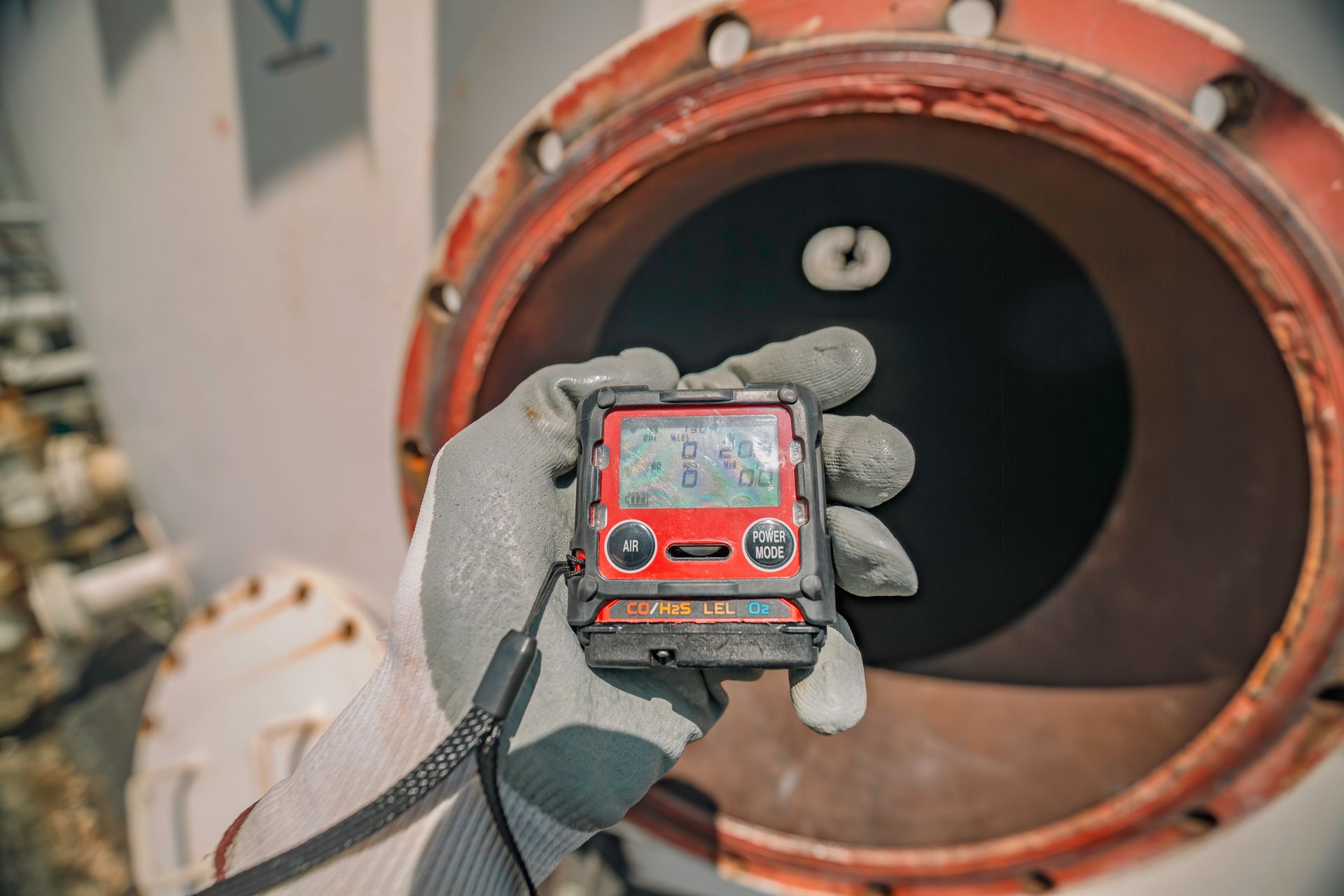 Gloved hand holding a gas detector in front of an open industrial pipe or tank opening. Gloved hand holding a gas detector in front of an open industrial pipe or tank opening.