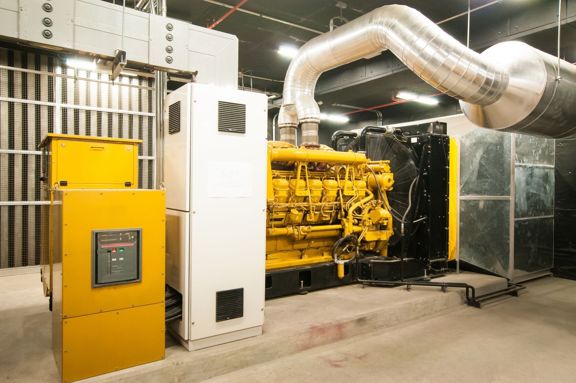 Yellow and black industrial generator in a room, connected to ductwork, with electrical panels.