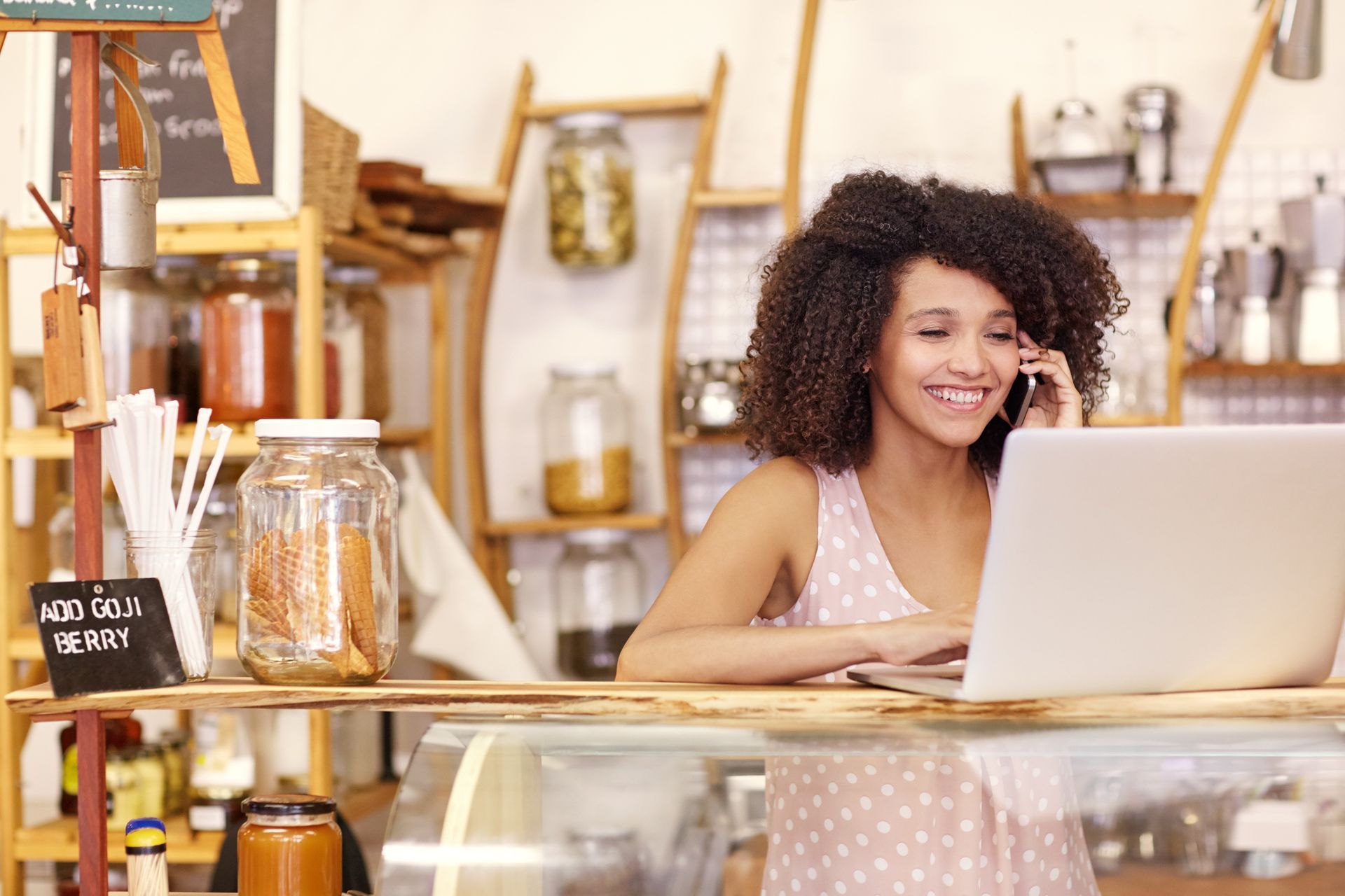 Woman smiling, on phone and using laptop at a cafe counter. Shelves of jars and goods are behind her.