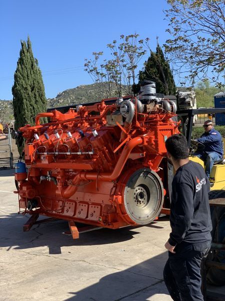 Large orange engine being moved outdoors by a forklift, two men observe on a sunny day.