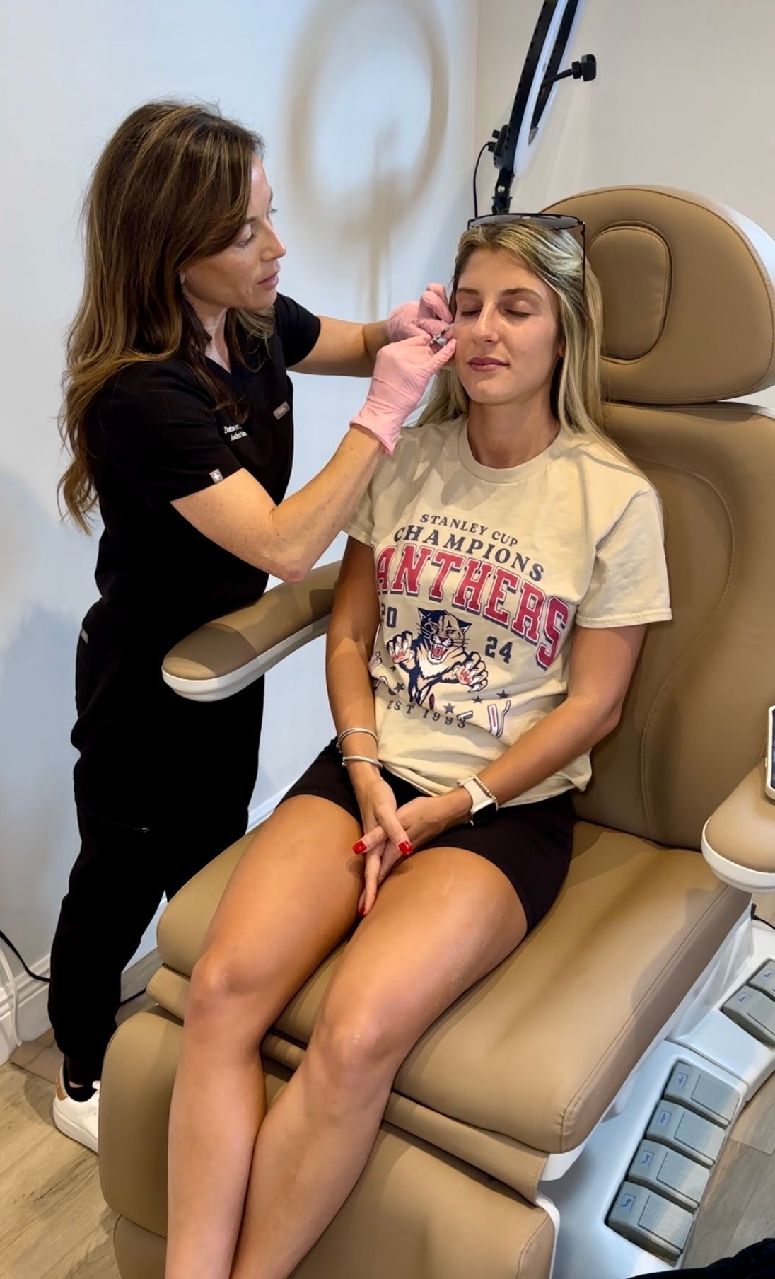 A practitioner wearing pink gloves performs a aesthetic injection on a patient sitting in a clinical chair.
